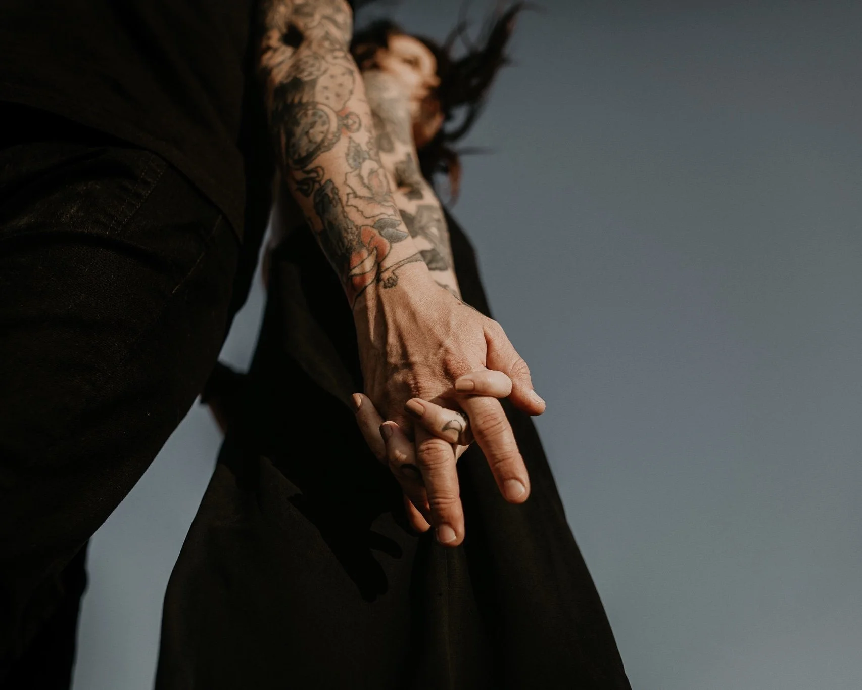 Nontraditional couple with tattoos on their arms and hands, wearing black clothing, holding hands against a plain sky background.