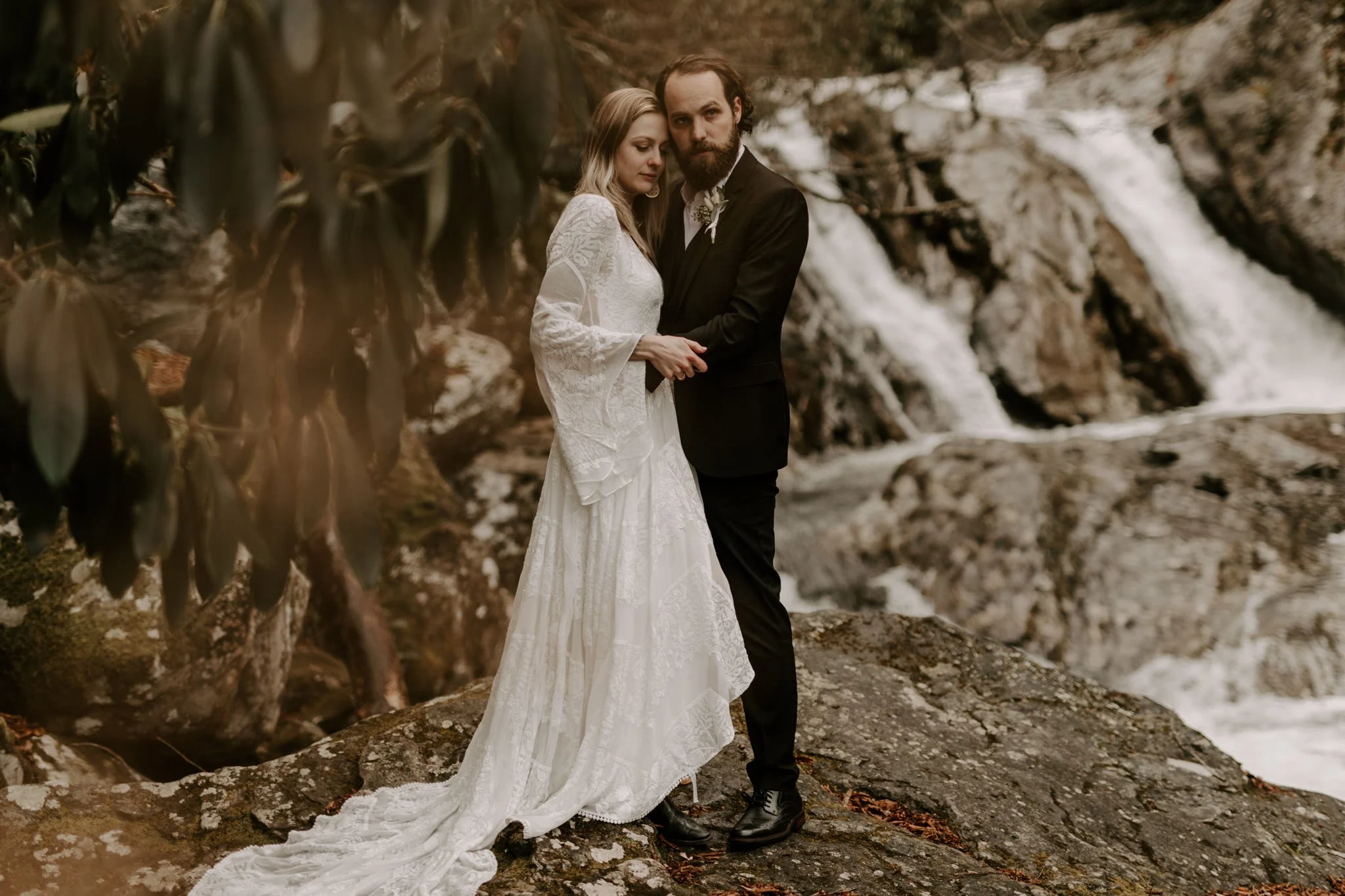 Bride and groom stand arm in arm on a rocky ledge with a waterfall behind them