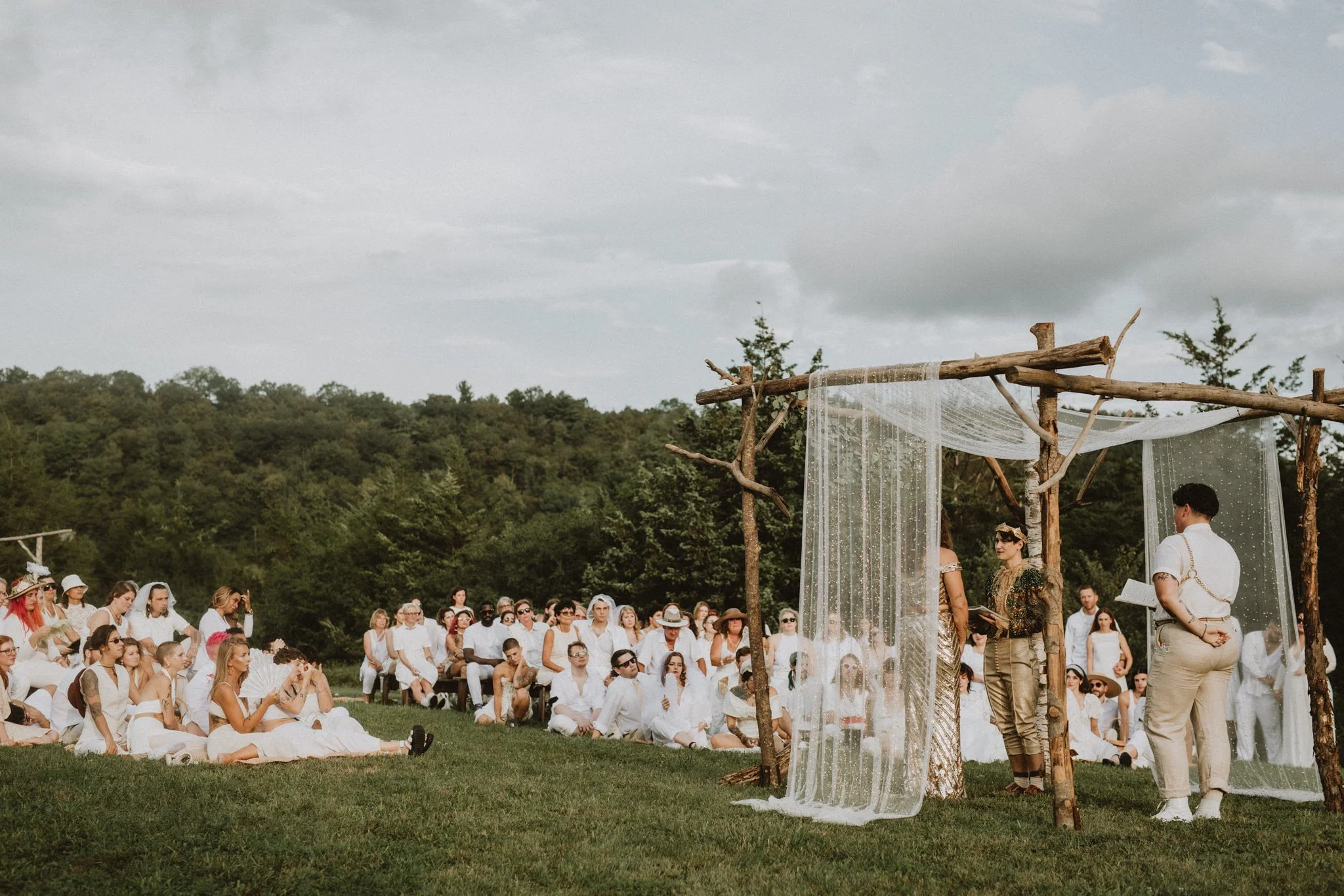 the brides stand under a wooden chuppa while their comment watches, all in white