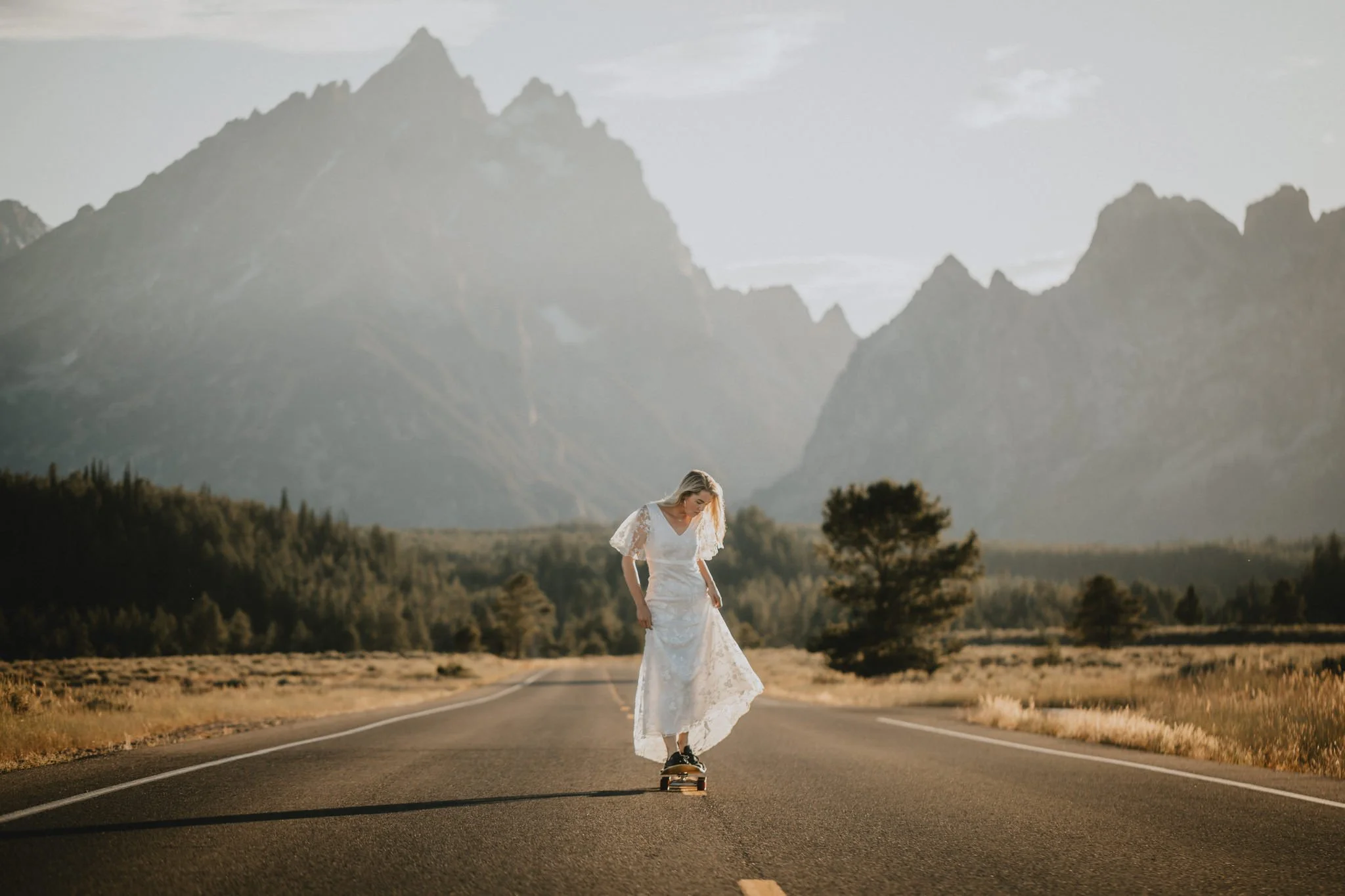 A bride in a wedding dress skateboarding on a deserted road with Grand Tetons in the background.