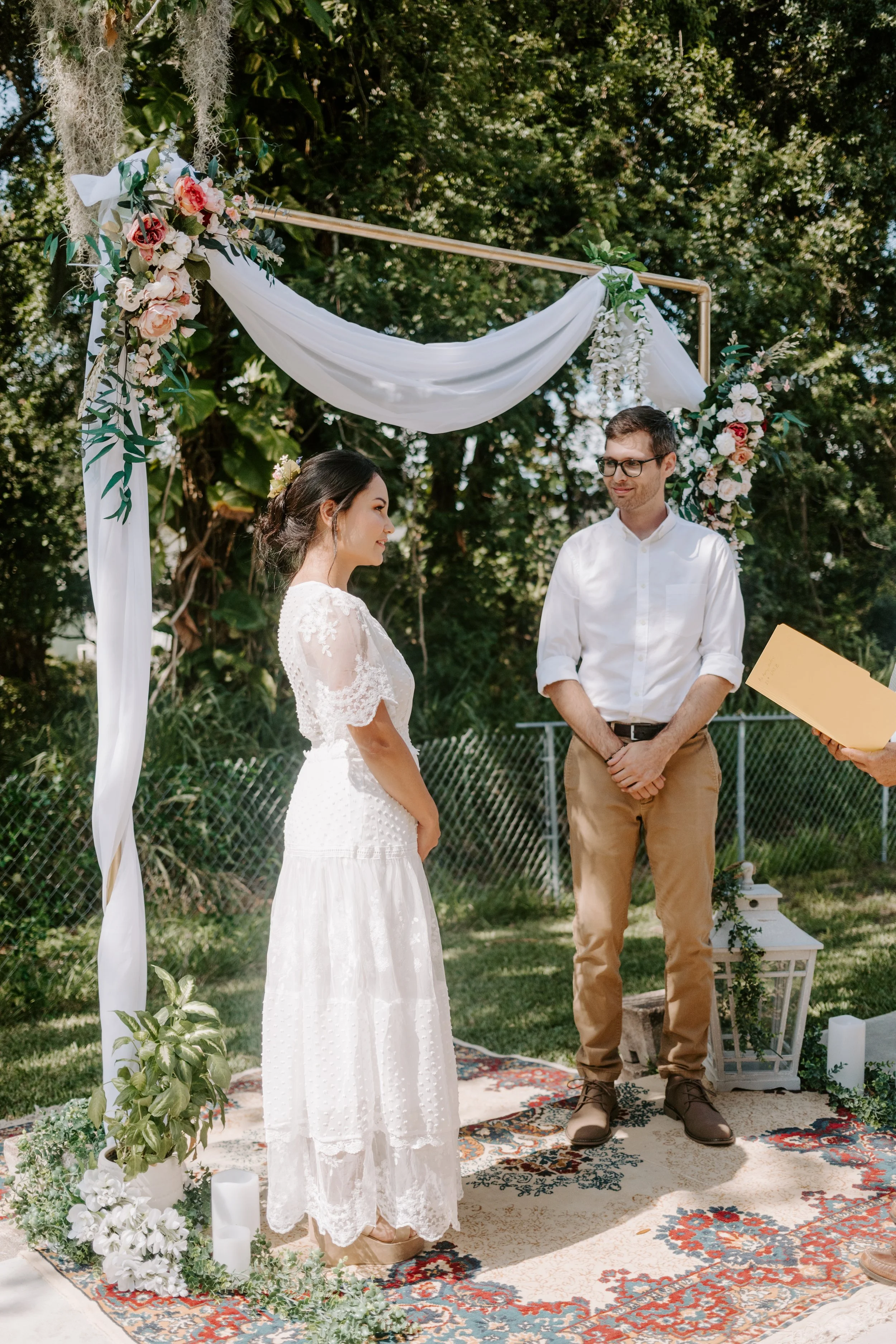 Bride speaks her vows to her father-in-law officiant as her groom smiles and watches, under their diy arch in their backyard