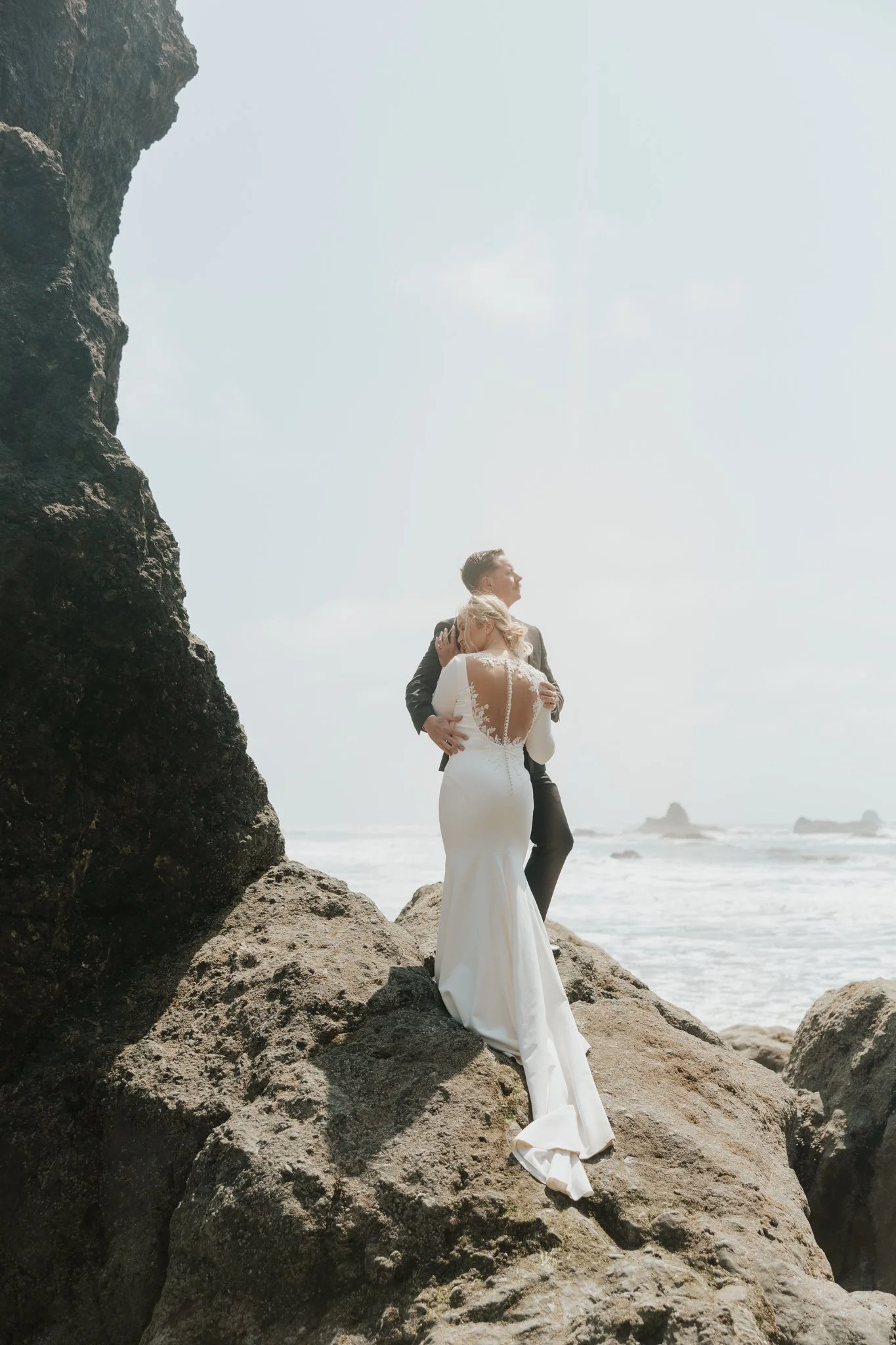 A bride and groom embracing on large rocks on Rialto Beach in Olympic National Park, at sunrise with a cloudy sky overhead.