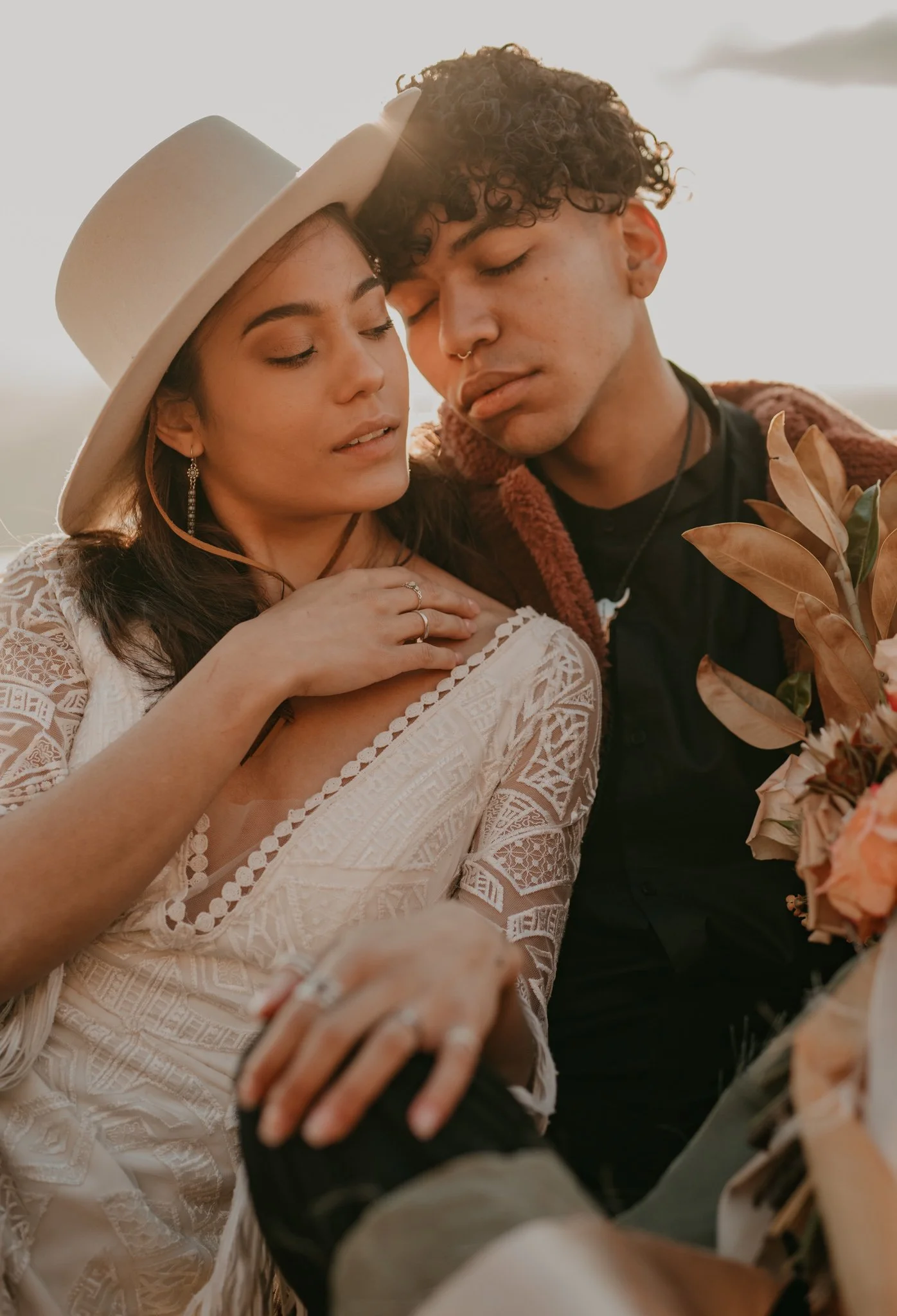 A boho wedding couple with curly hair are close, eyes closed, sharing an intimate moment at Dead Horse Point State Park. The woman wears a wide-brimmed hat and a lace dress, and the man holds a bouquet of flowers.
