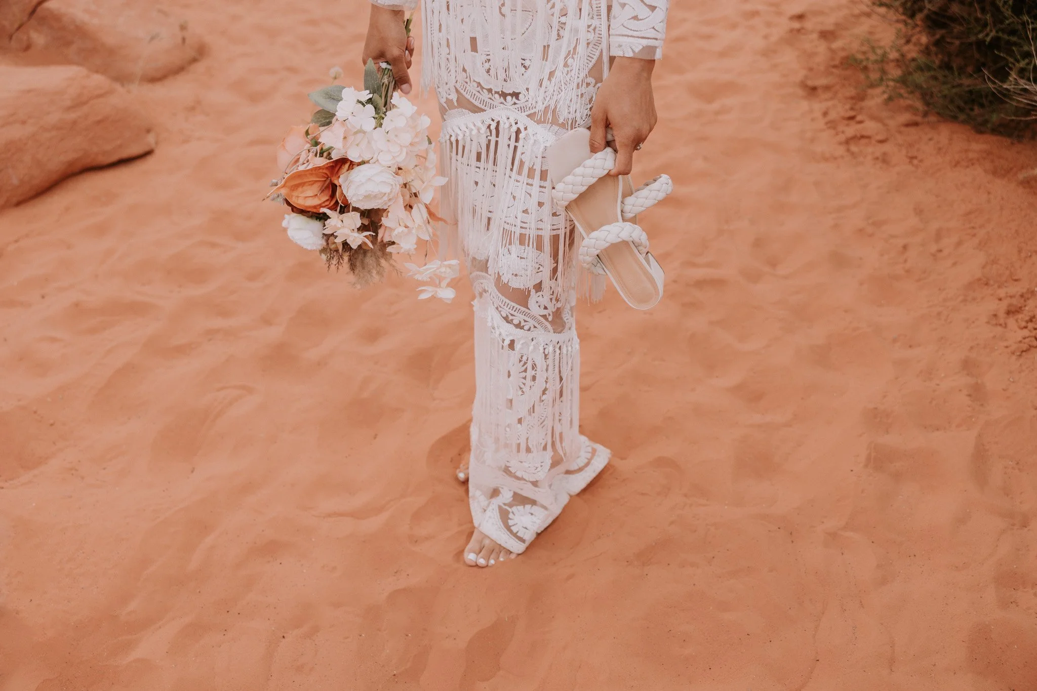 A woman wearing a white lace wedding dress holding a bouquet of flowers and a pair of white shoes with braided straps on a sandy surface in Valley of Fire.