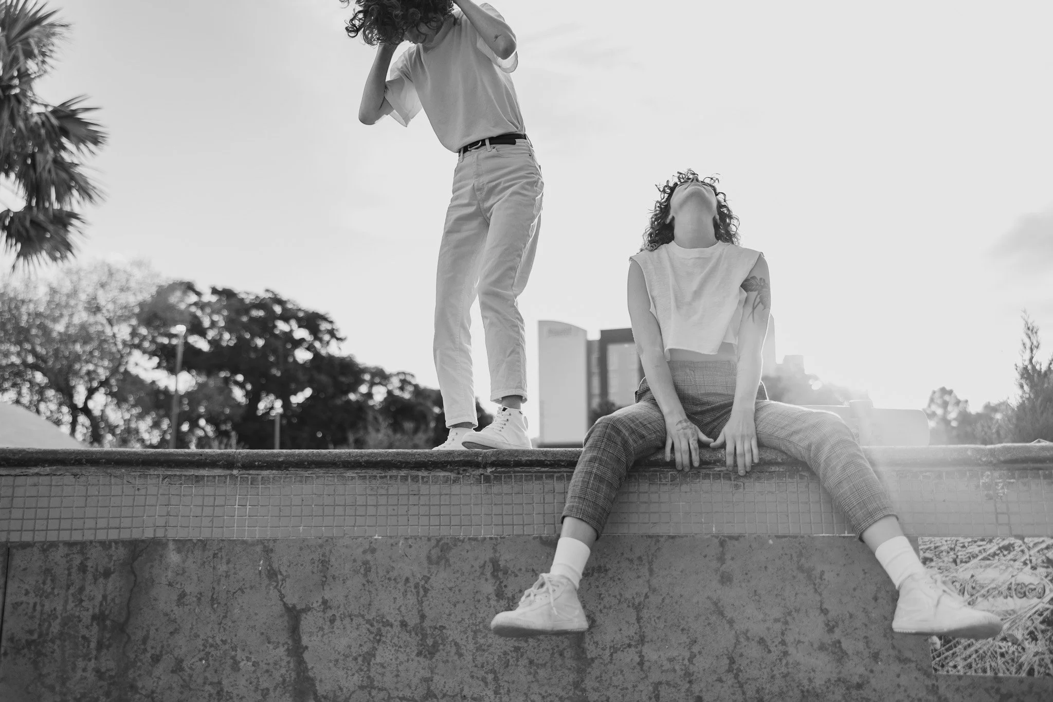 Queer couple with curly hair, one standing and one sitting on a ledge of a skate park bowl, outdoors in St. Pete, Florida, captured in black and white.