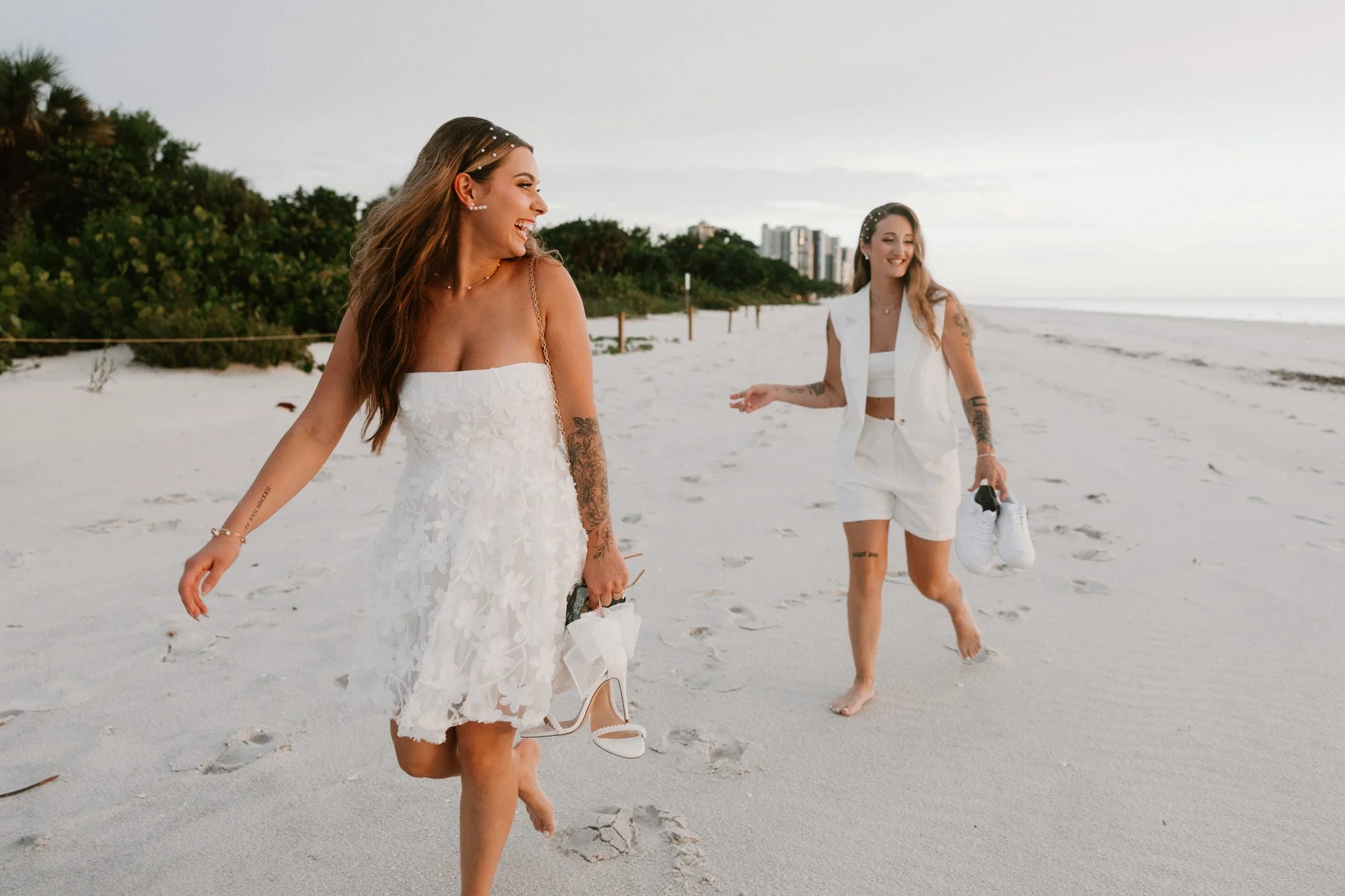 Two women walking barefoot on a beach, smiling and holding their shoes, with one wearing a white wedding dress and the other in a white sleeveless vest and shorts, in a bright, scenic beach setting with greenery and buildings in the background.