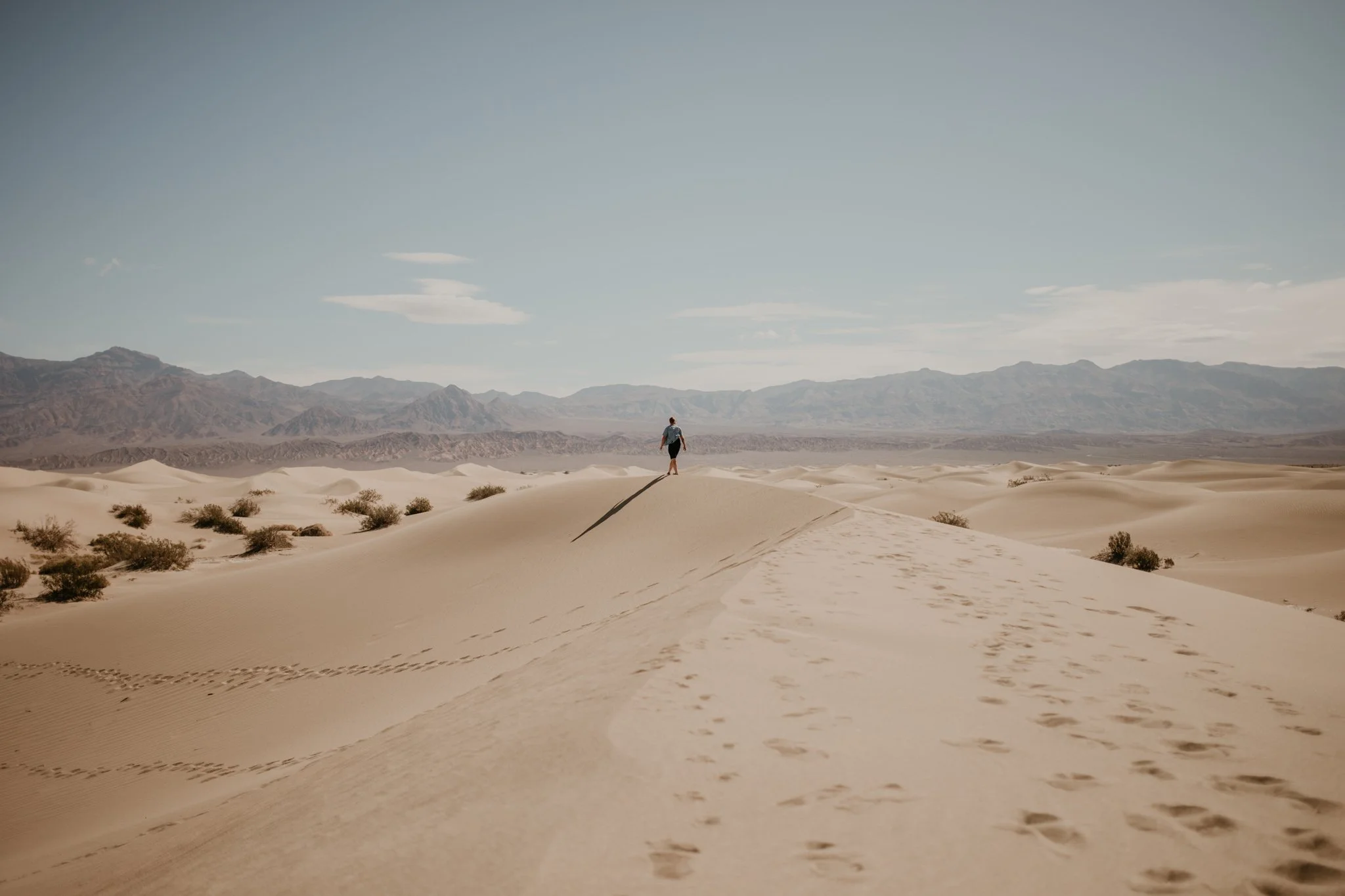 Corey of Going Home Co. walking alone on sand dunes at Death Valley National Park, with mountains in the background and a clear sky.