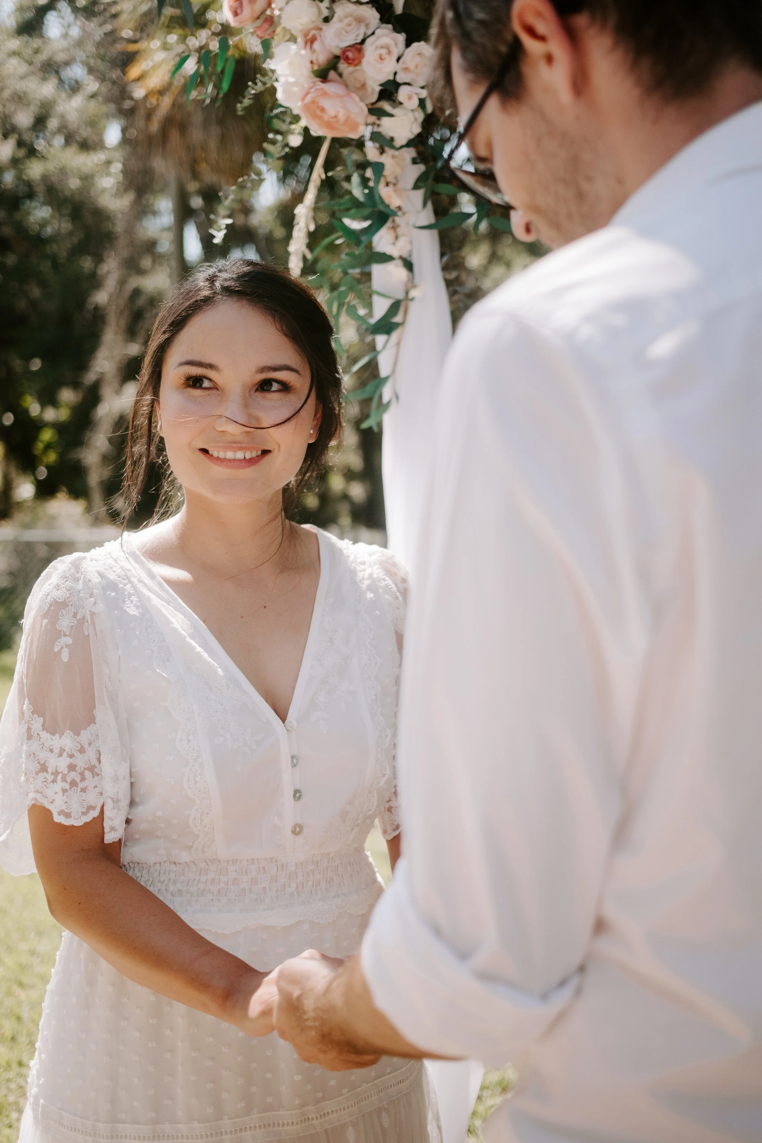 Bride smiles while standing hand-in-hand wit her groom, under a floral arch