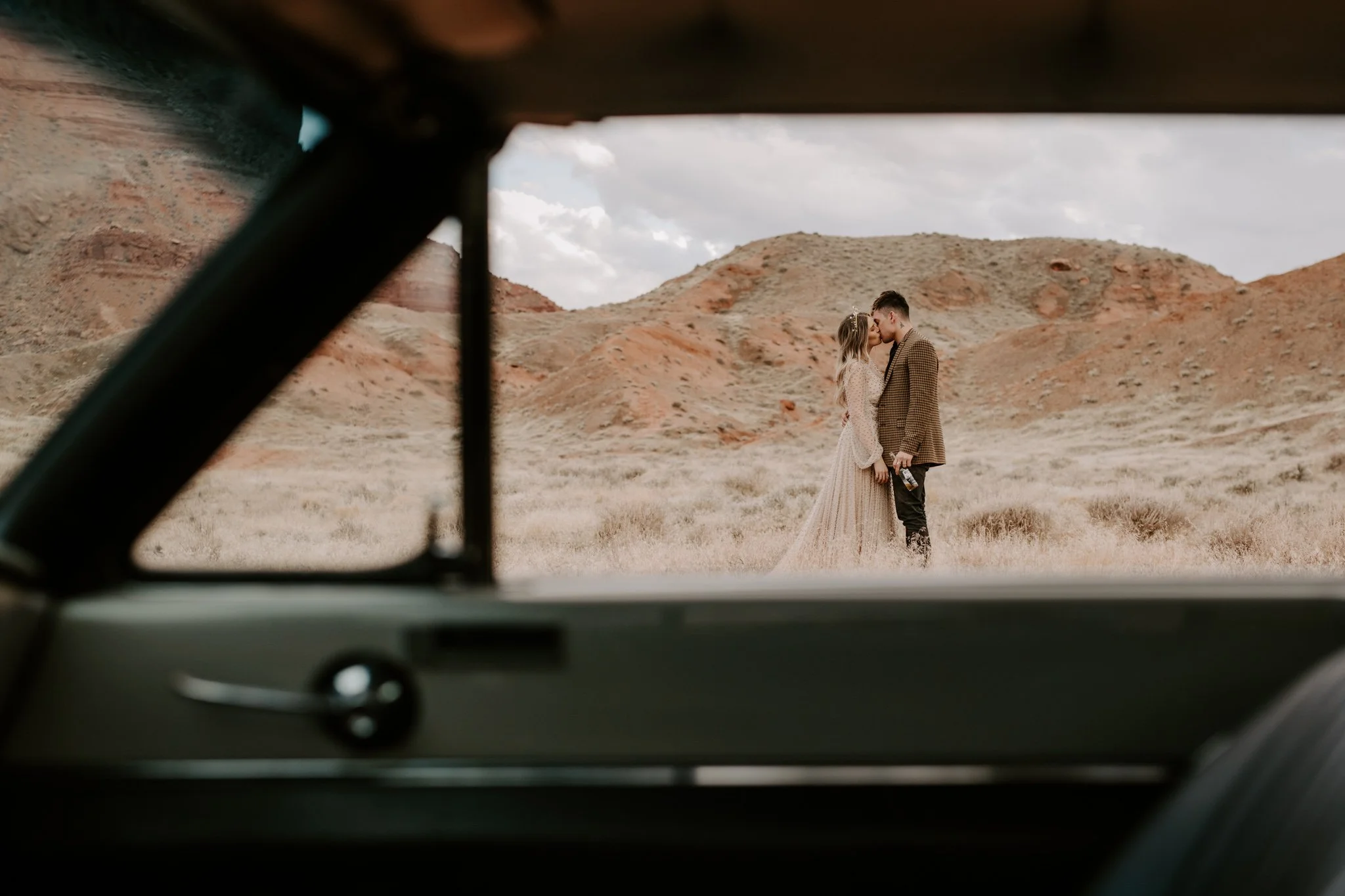 A tattooed couple is kissing in a desert landscape, seen through the window of a classic car.