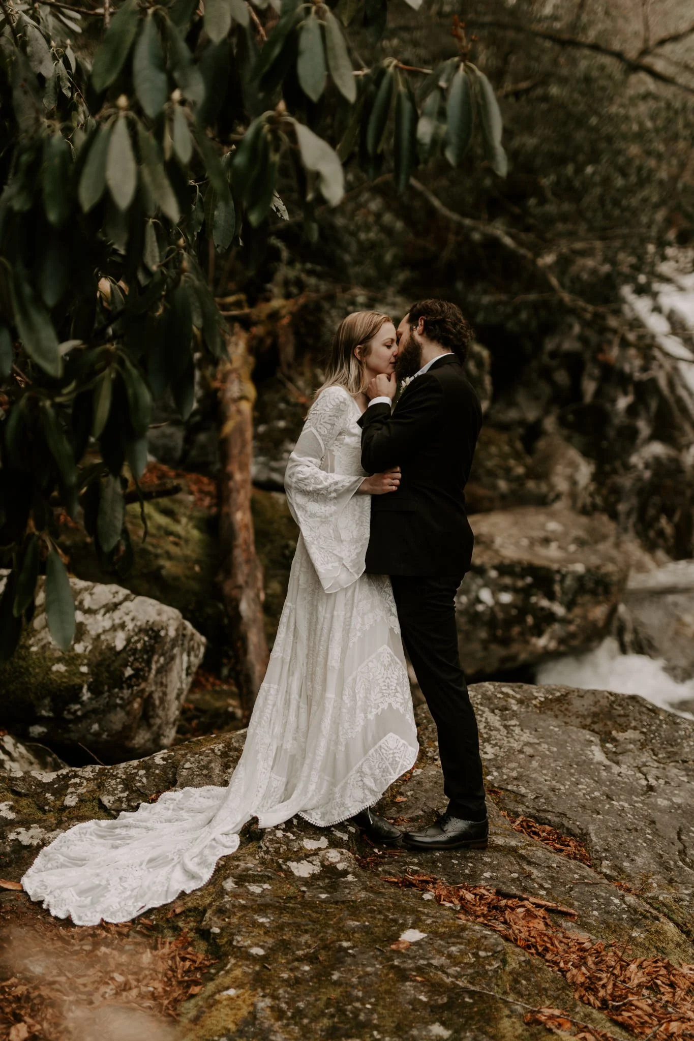 Bride and groom share a kiss on a rocky ledge next to a waterfall in Pisgah national forest