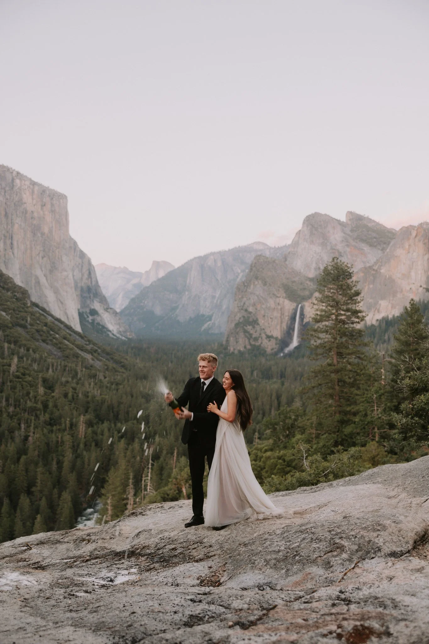 A bride and groom celebrating their elopement on a rocky ledge in Yosemite National Park, with the groom opening a champagne bottle, surrounded by tall trees and foggy Valley View in the background.