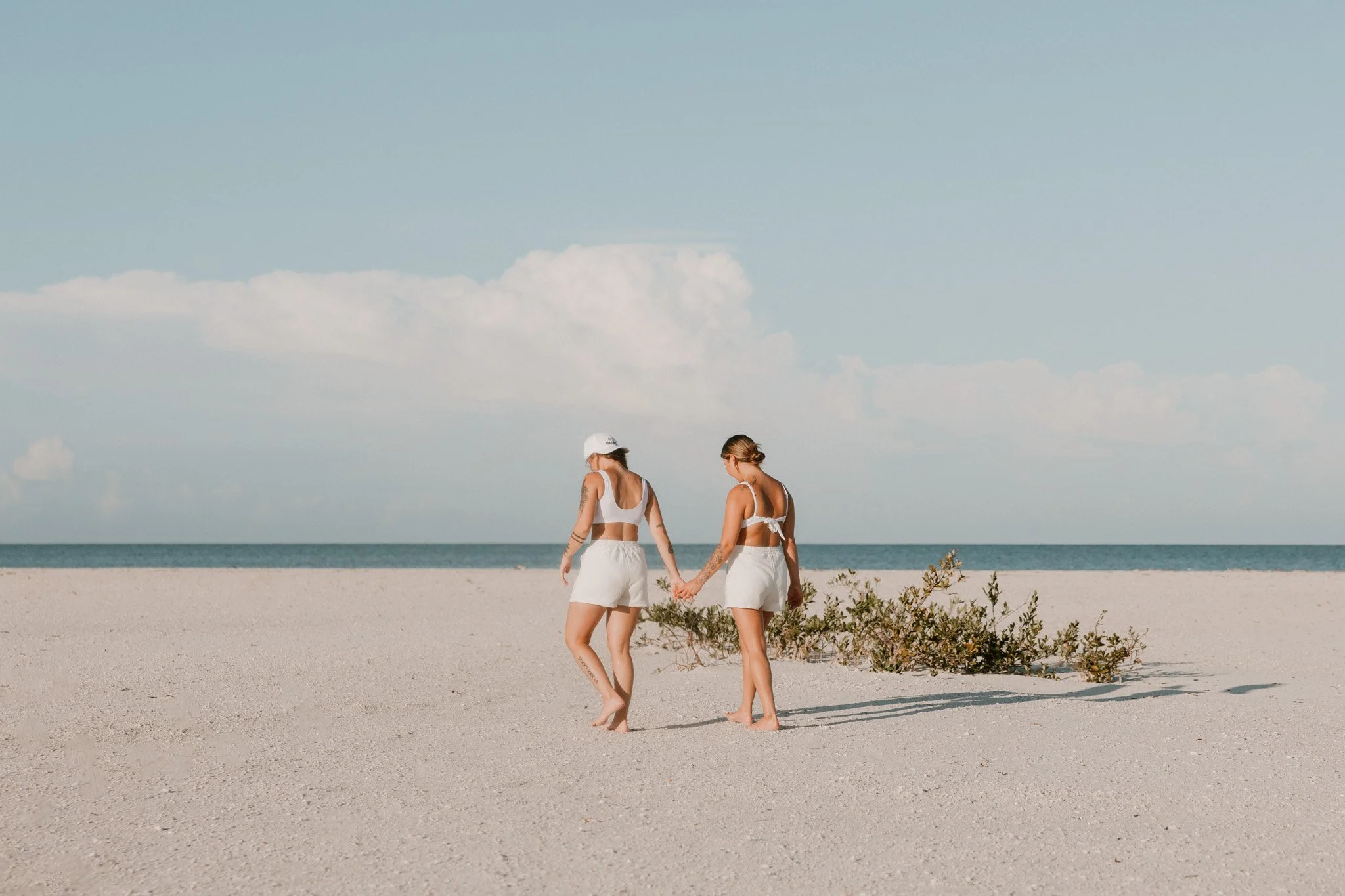 Queer couple in white outfits holding hands and walking on a sandy beach with a blue sky and ocean in the background.