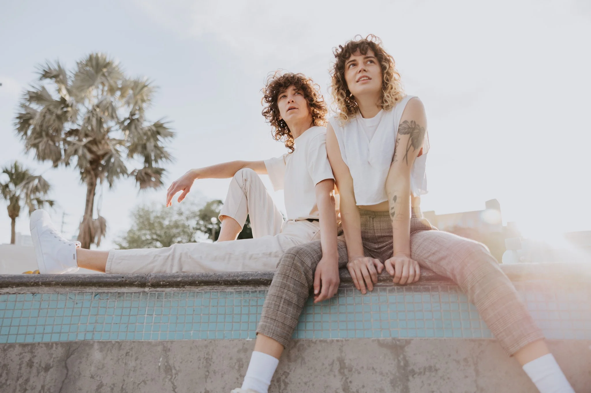 Two young queer women with curly hair sitting on the edge of a skate park bowl, looking up at the sky, with palm trees and a clear sky in the background.