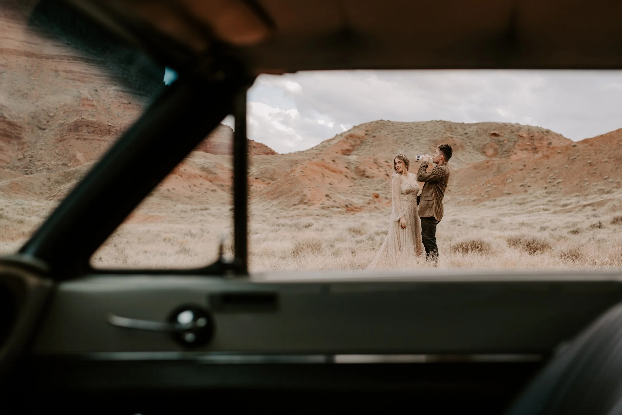 Couple shares a beer while hiking around Moab with their retro car in the foreground