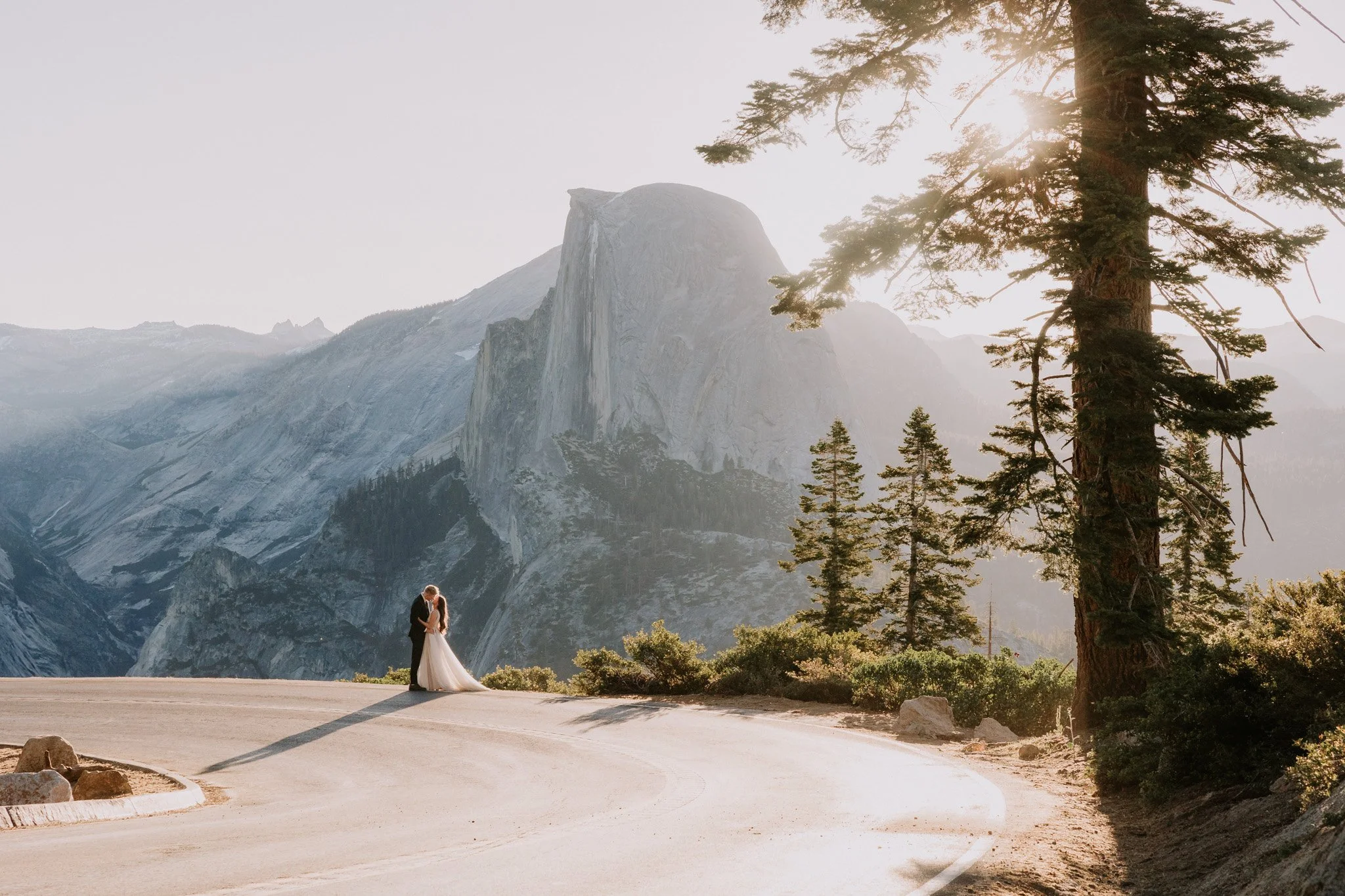 A bride and groom hold each other and kiss on a winding mountain road in Yosemite with Half Dome in the background during late morning.