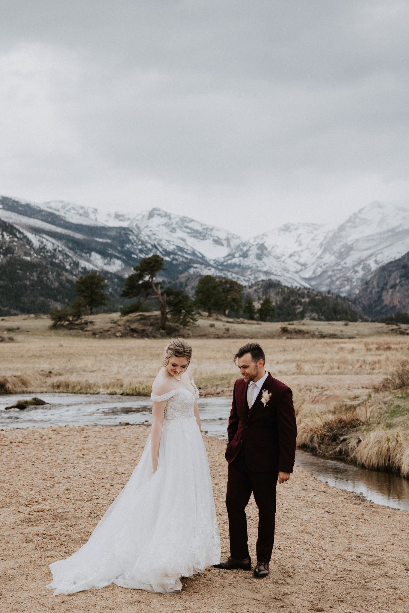 A bride and groom standing together in Rocky Mountain National Park with snow-capped peaks, a river, and grass fields in the background.