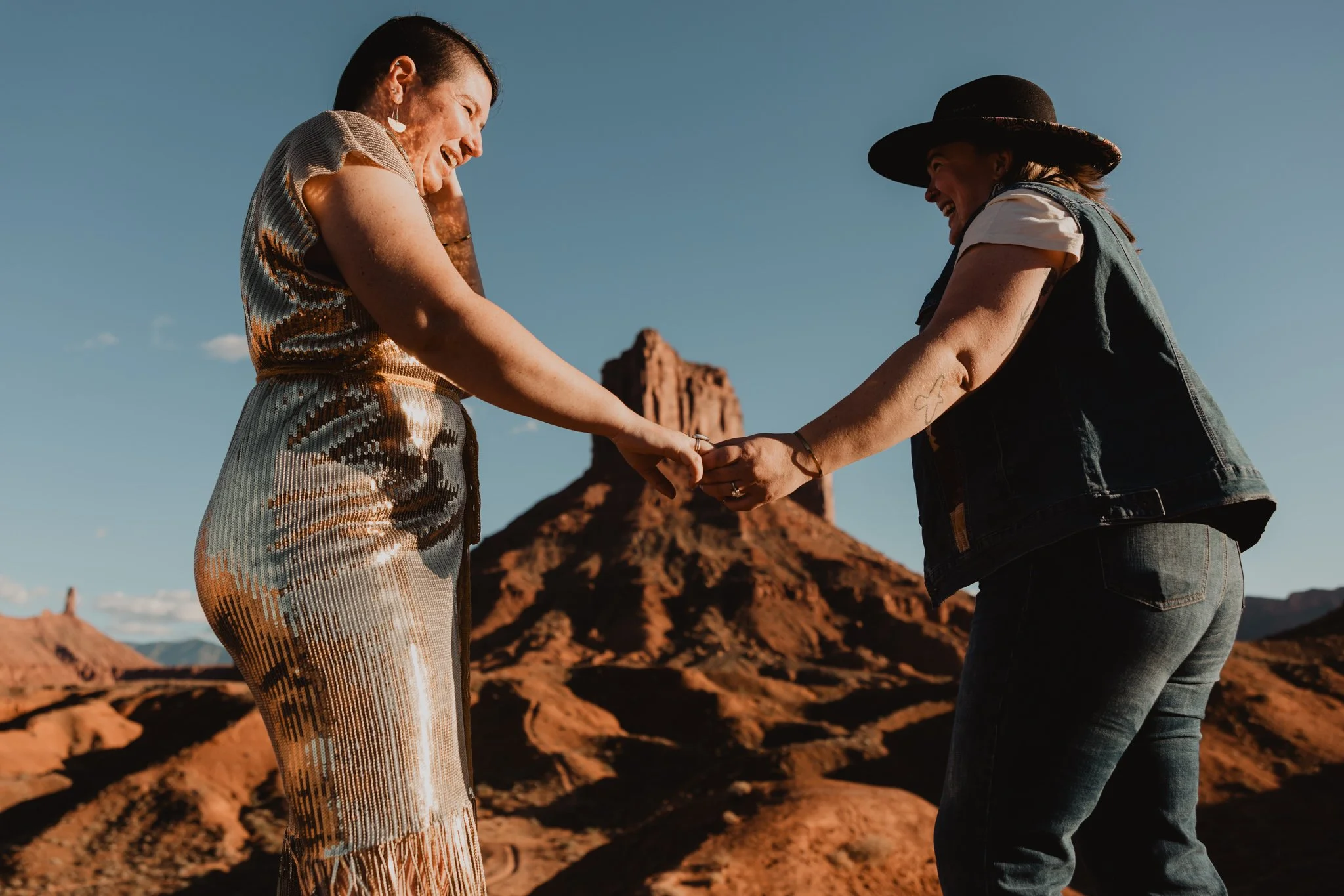 A queer couple at Ida Gulch in Moab, holding hands and dancing in front of a mesa
