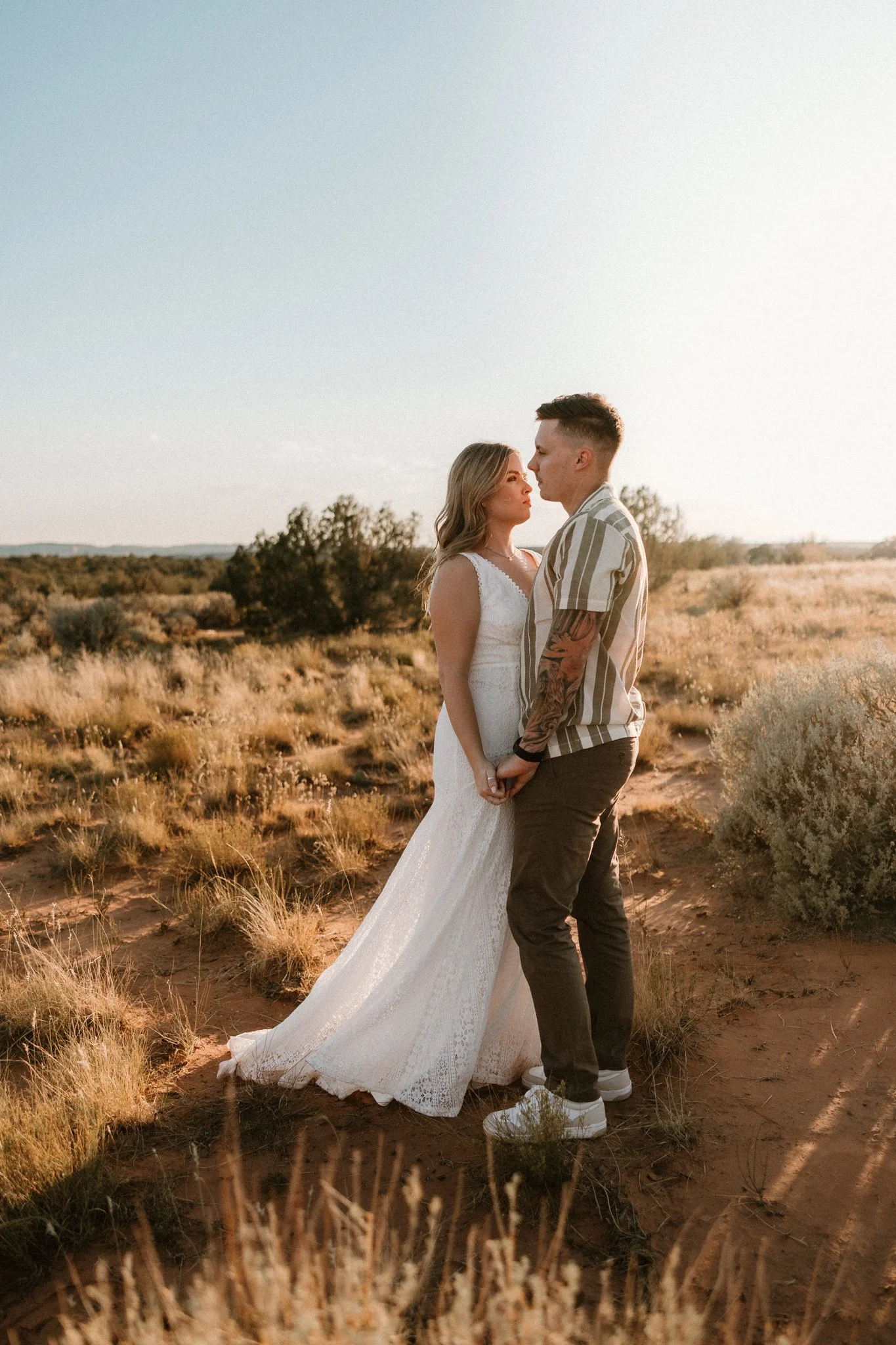A tattooed couple stand close together and hold hands in the Moab desert with the sun setting behind them
