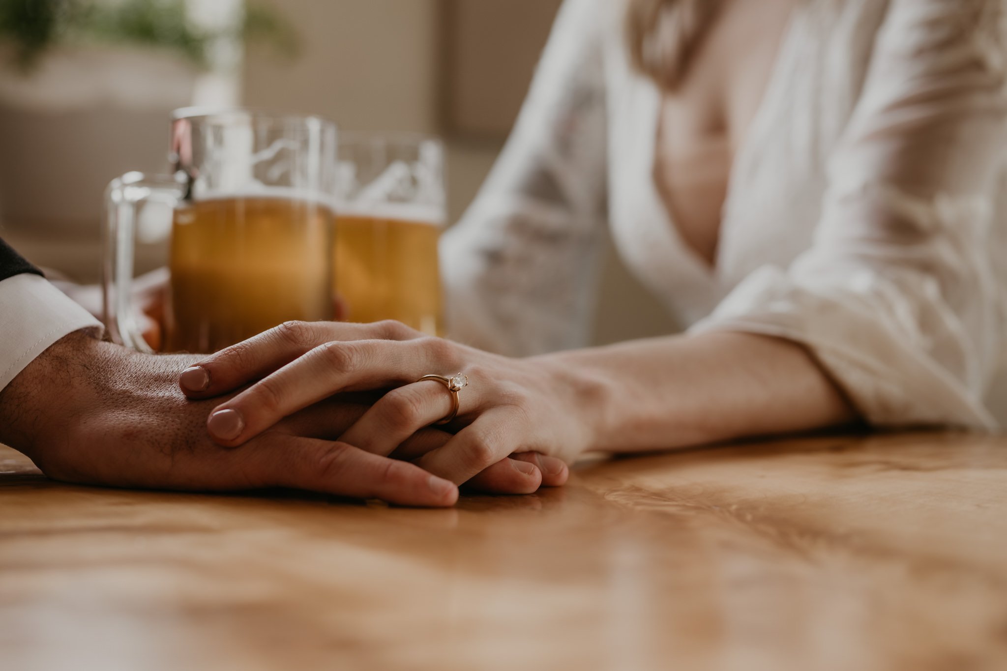 A brides hand sit ontop of her grooms hand, displaying her engagement ring, beers sitting on the table next to them