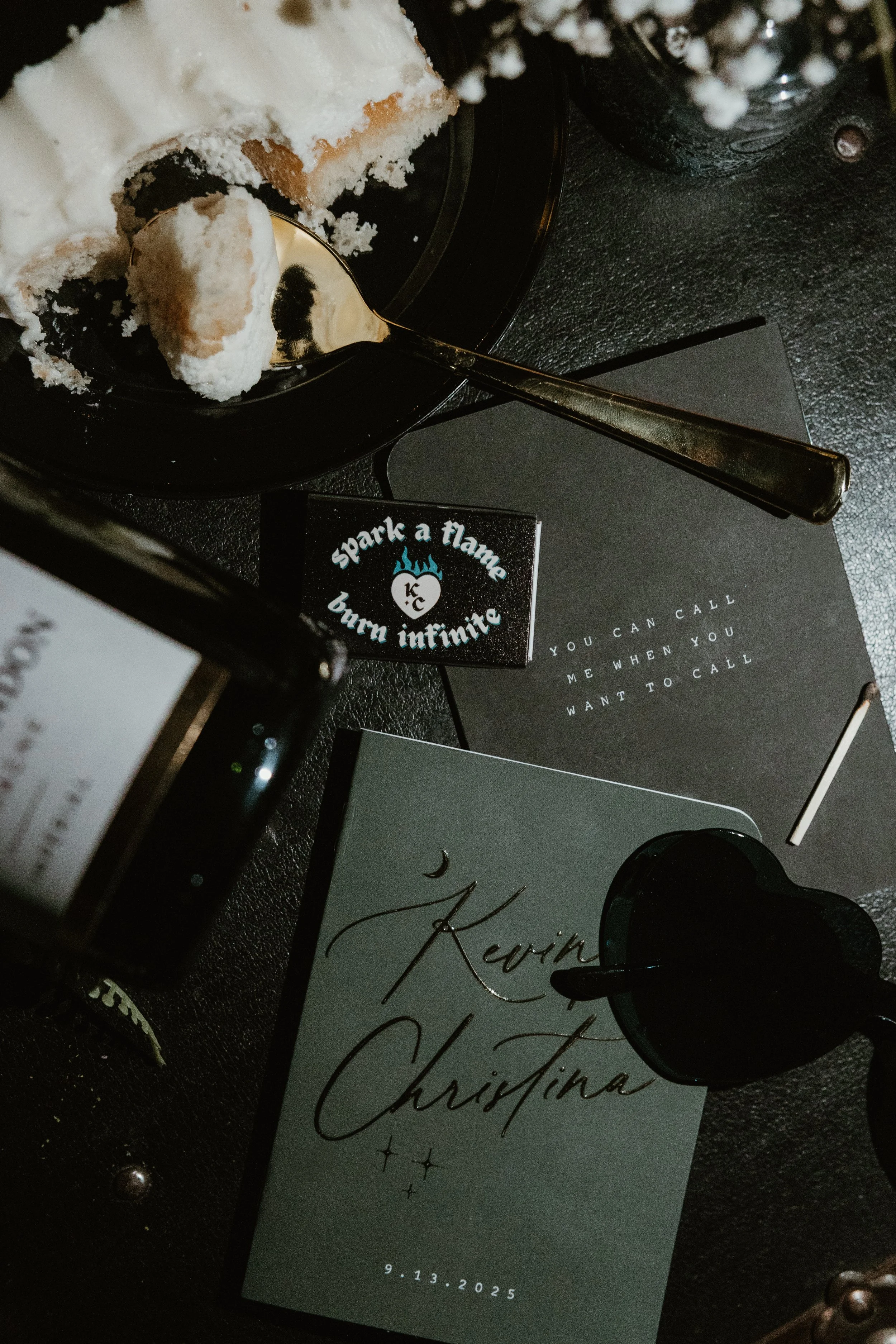A black table displays a cake slice with white frosting, a spoon, a black card with white and blue text, a black notebook with white text, a black glass with a heart-shaped handle, a wedding invitation, and a black bottle partially visible.