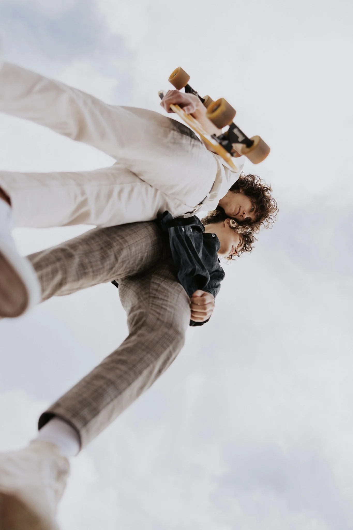 Queer couple standing on the edge of a skate park bowl, one holding a skateboard, viewed from a low angle against a cloudy sky.