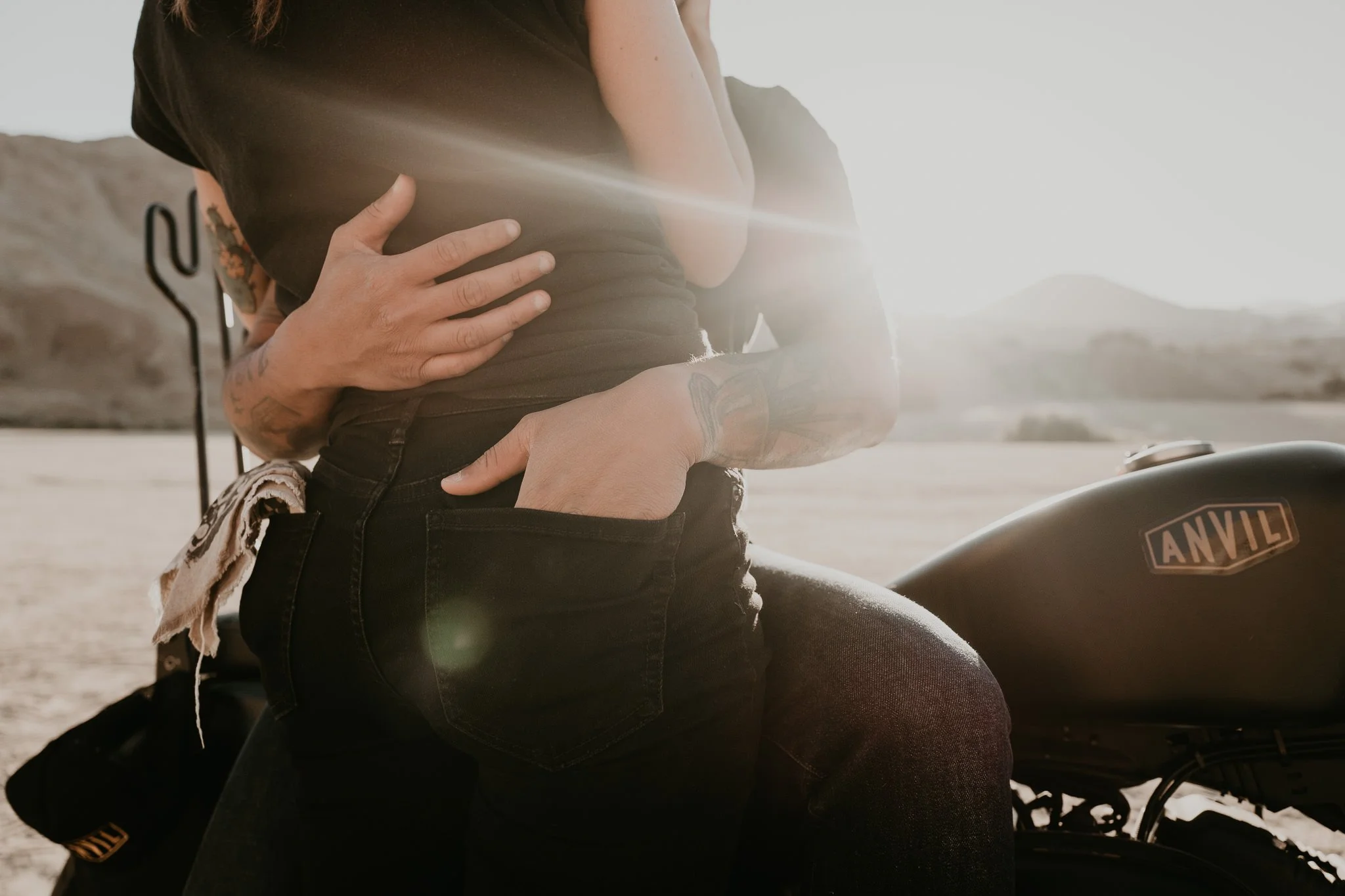 Close up of a tattooed couple sitting on a motorcycle with his hand in her back pocket, outdoors in California at sunset. The motorcycle has a visible 'Anvil' logo on the fuel tank.