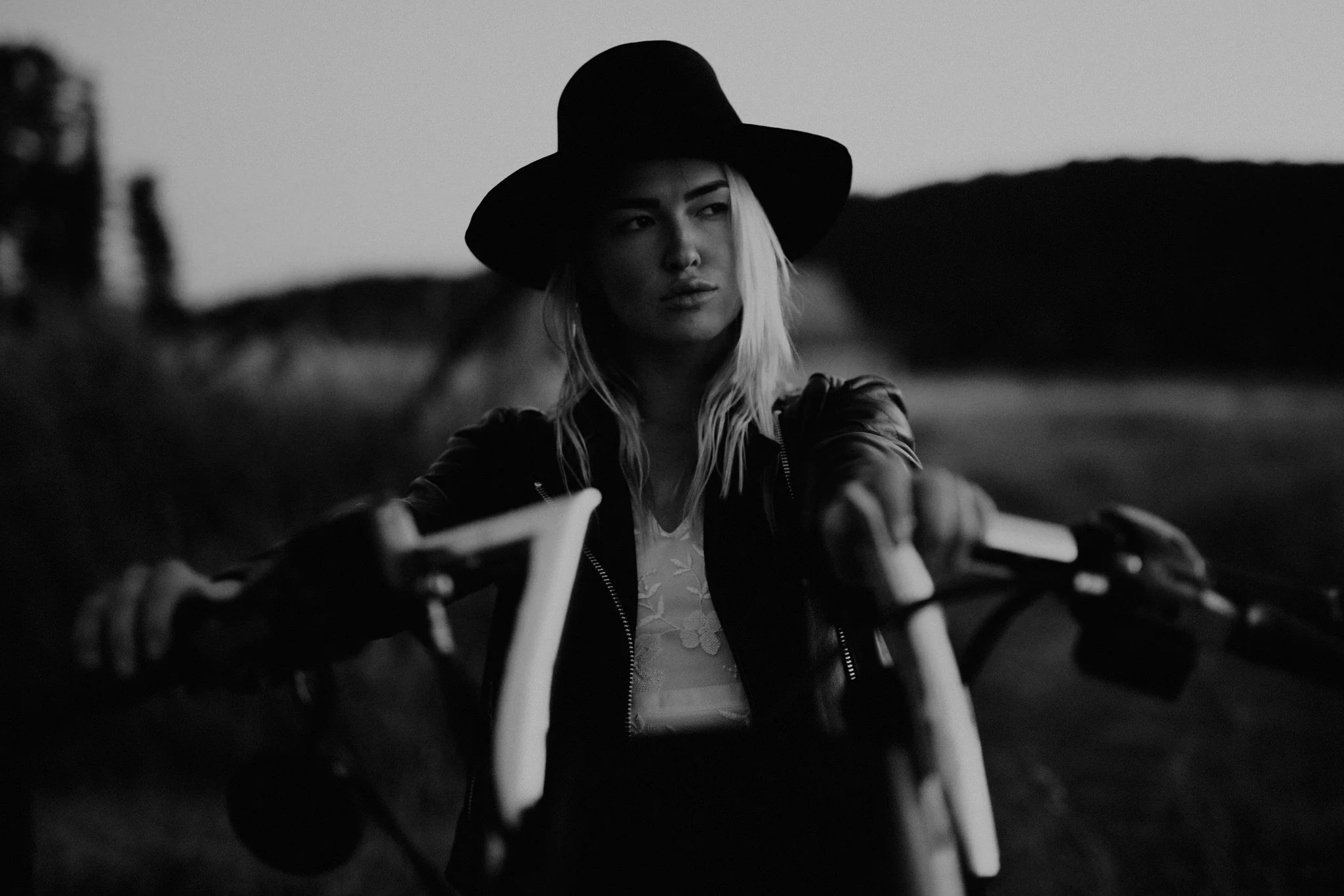 A woman with blonde hair wearing a large black hat, wedding dress and a leather jacket, sitting on a motorcycle and looking to the side outdoors during dusk.