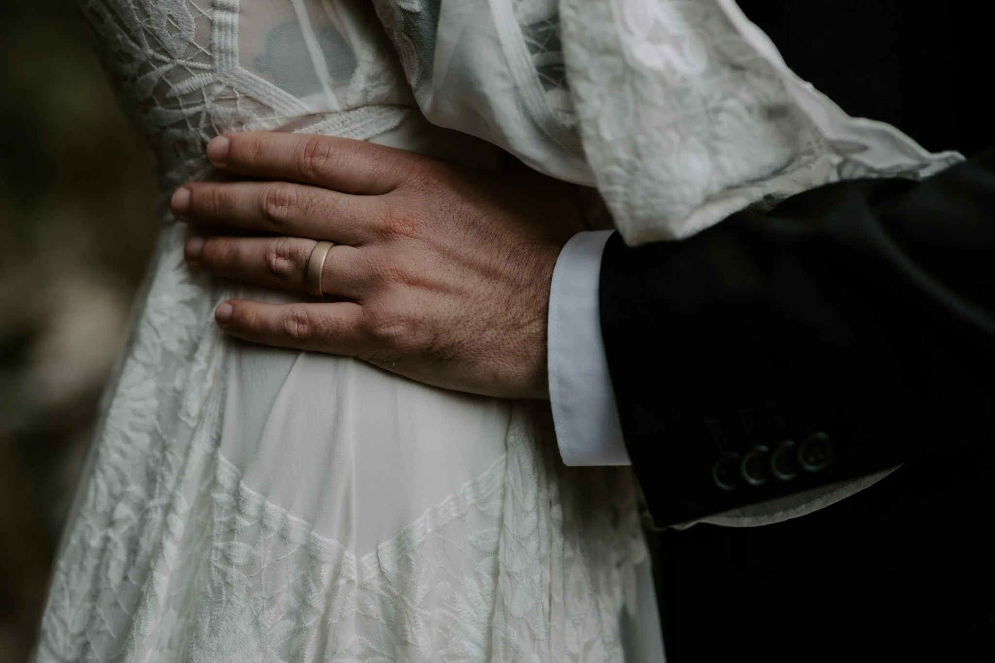 A closeup of a grooms hand and ring, as it holds his brides waist