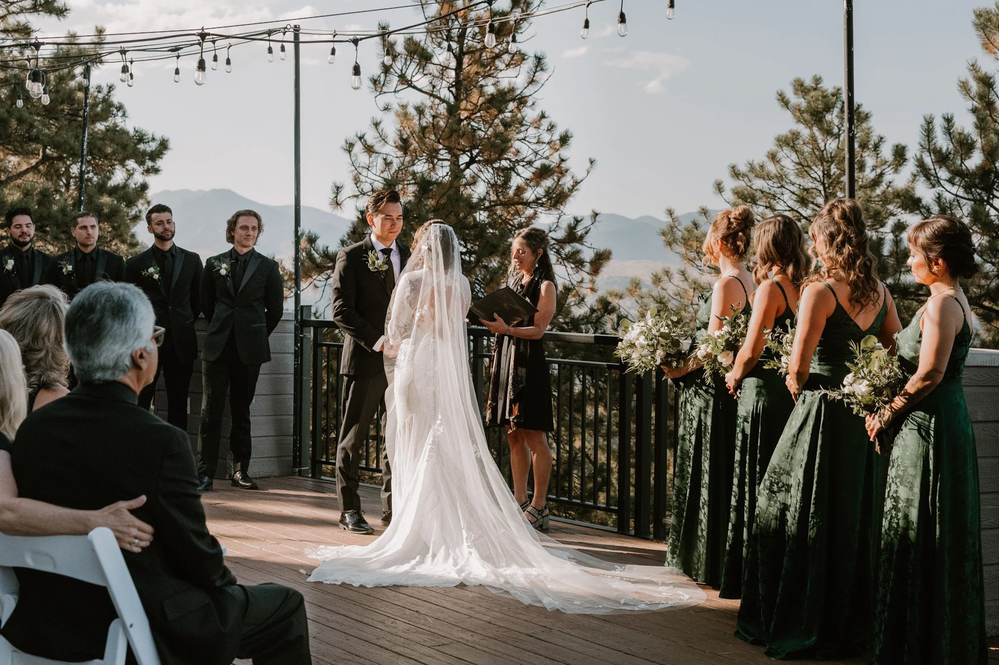 A wedding ceremony with a bride and groom exchanging vows on a wooden deck outdoor setting with mountain views in Colorado, surrounded by bridesmaids in green dresses holding bouquets and groomsmen in black suits, under string lights.