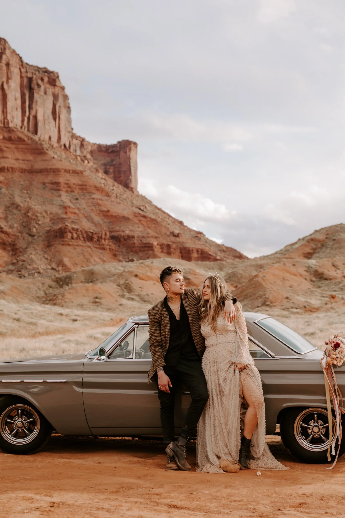 Nontraditional wedding couple stands against their retro car in the Moab desert at sunset