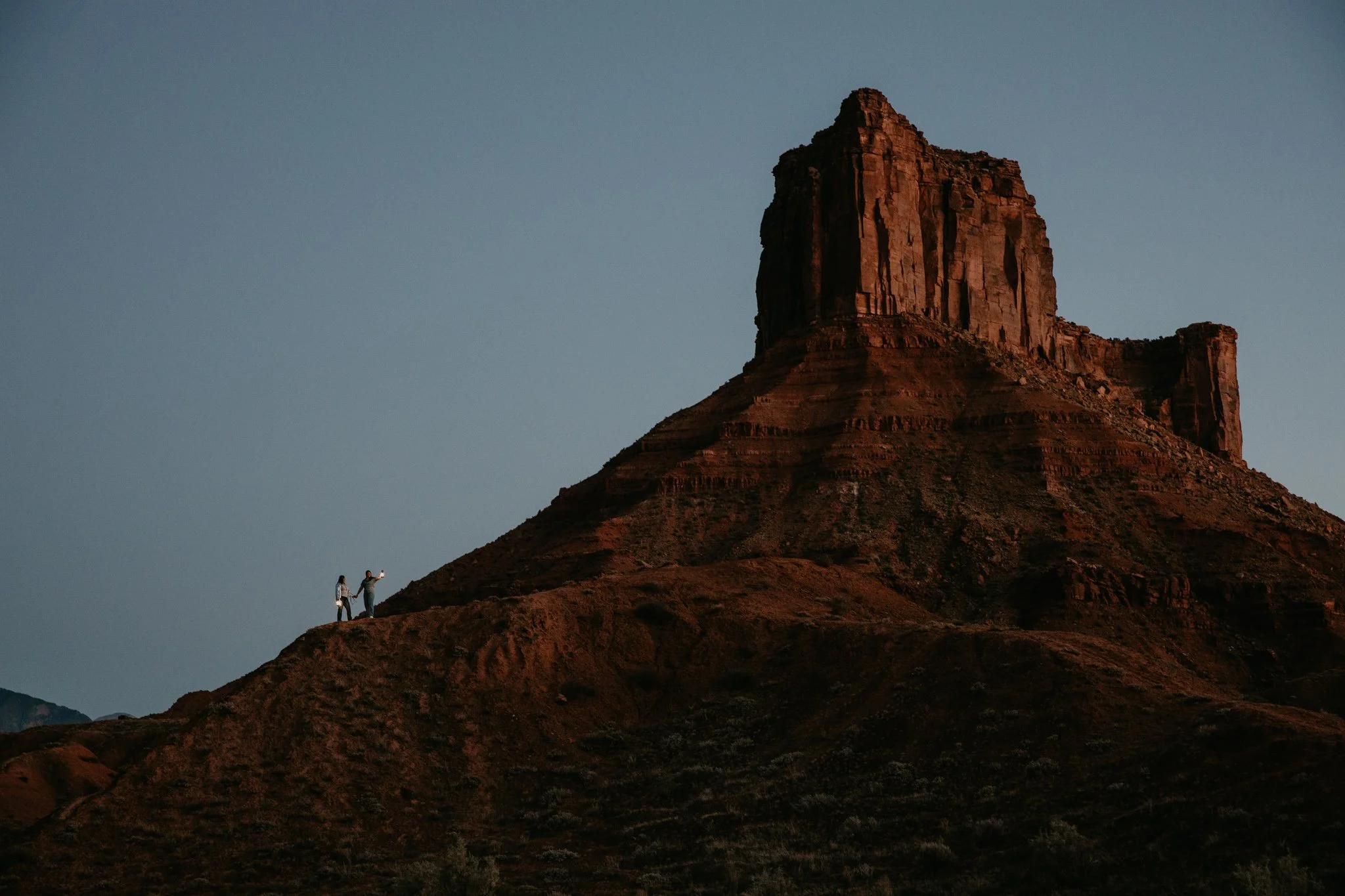 Queer couple standing on a small hilltop, holding lanterns in Moab with a large rock mesa formation in the background during dusk.