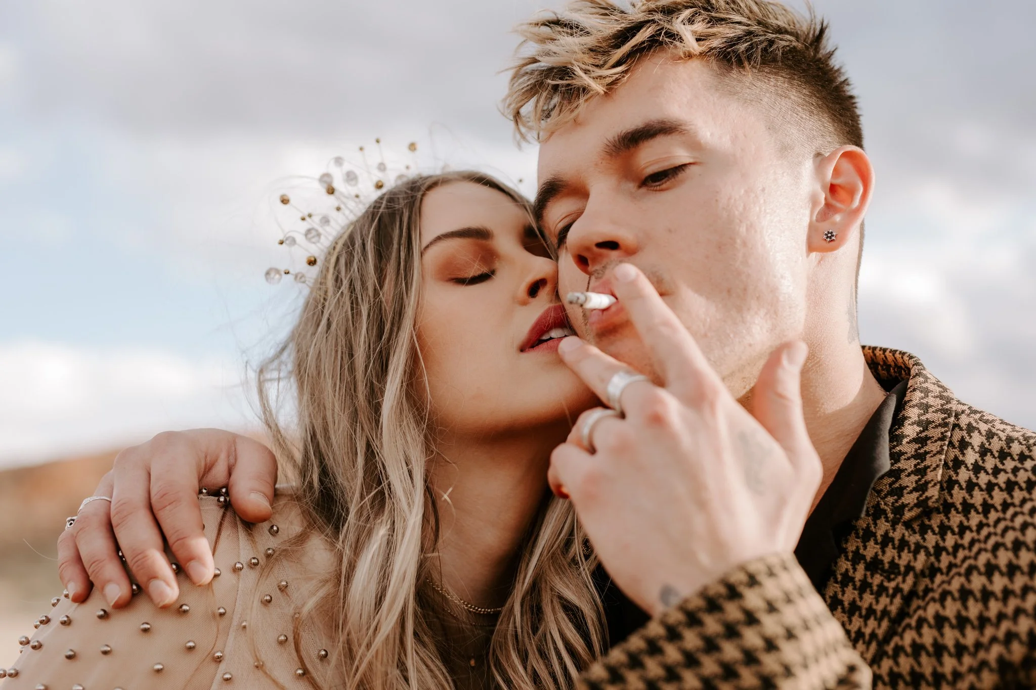 A young nontraditional couple close together in Moab, with the woman wearing a pearl-studded wedding dress, and the tattooed man wearing a houndstooth jacket. They're sharing a moment, with the man holding a cigarette.