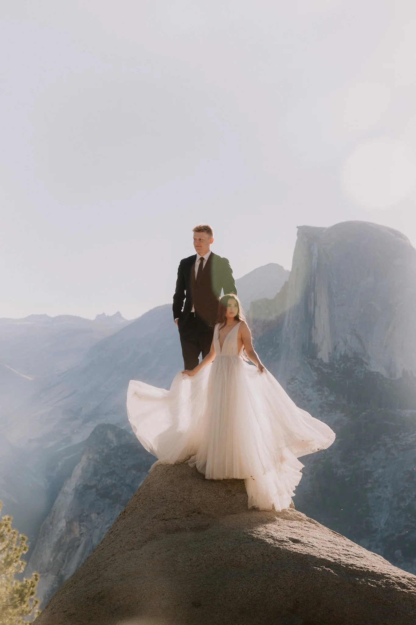 A groom in a black suit and a bride in a white wedding dress standing on a large rock with Half Dome in the background.