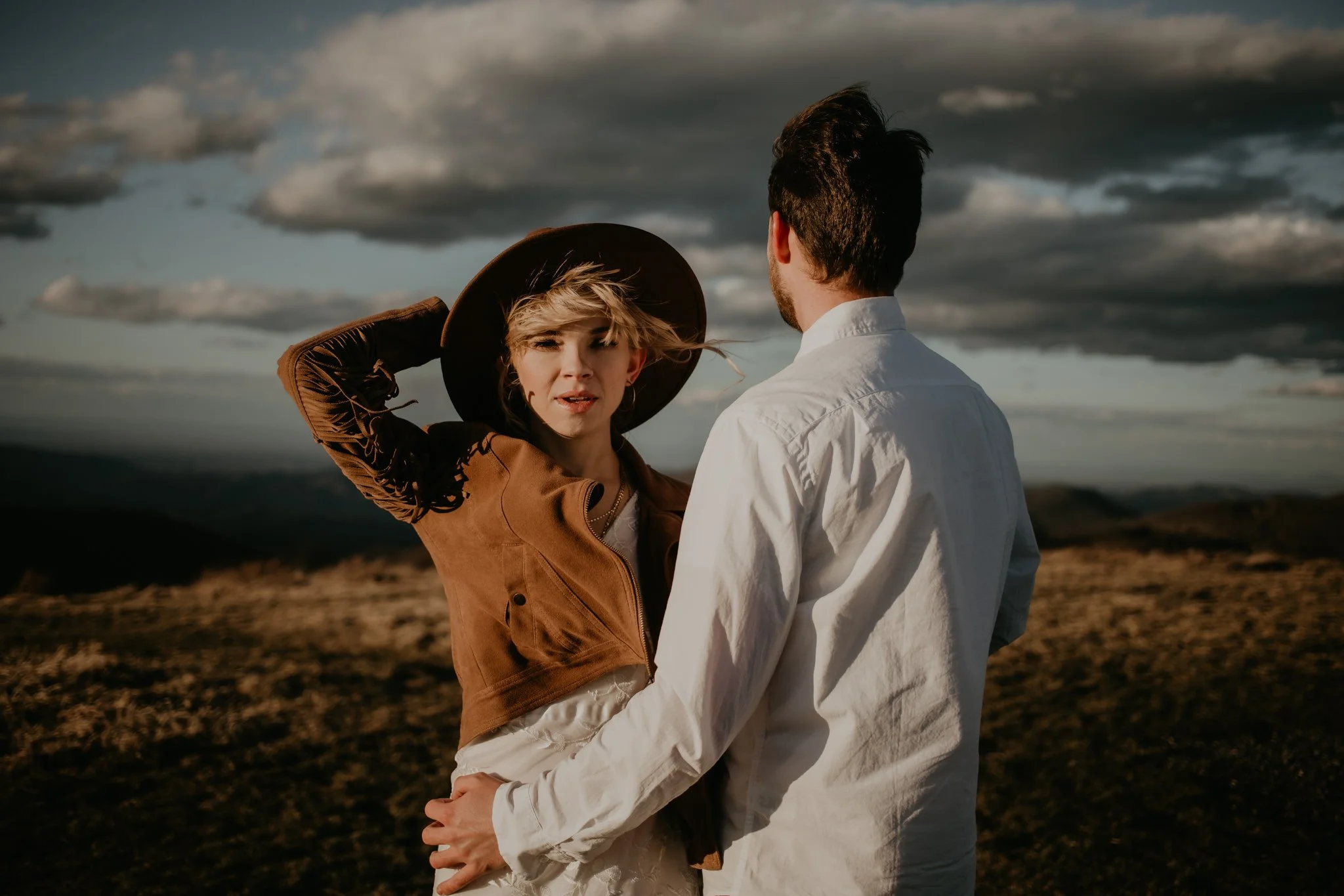 A young bride in a brown fringe jacket and wide-brimmed hat looks at her groom in a white shirt, standing in a landscape with dark clouds and hills in the background.