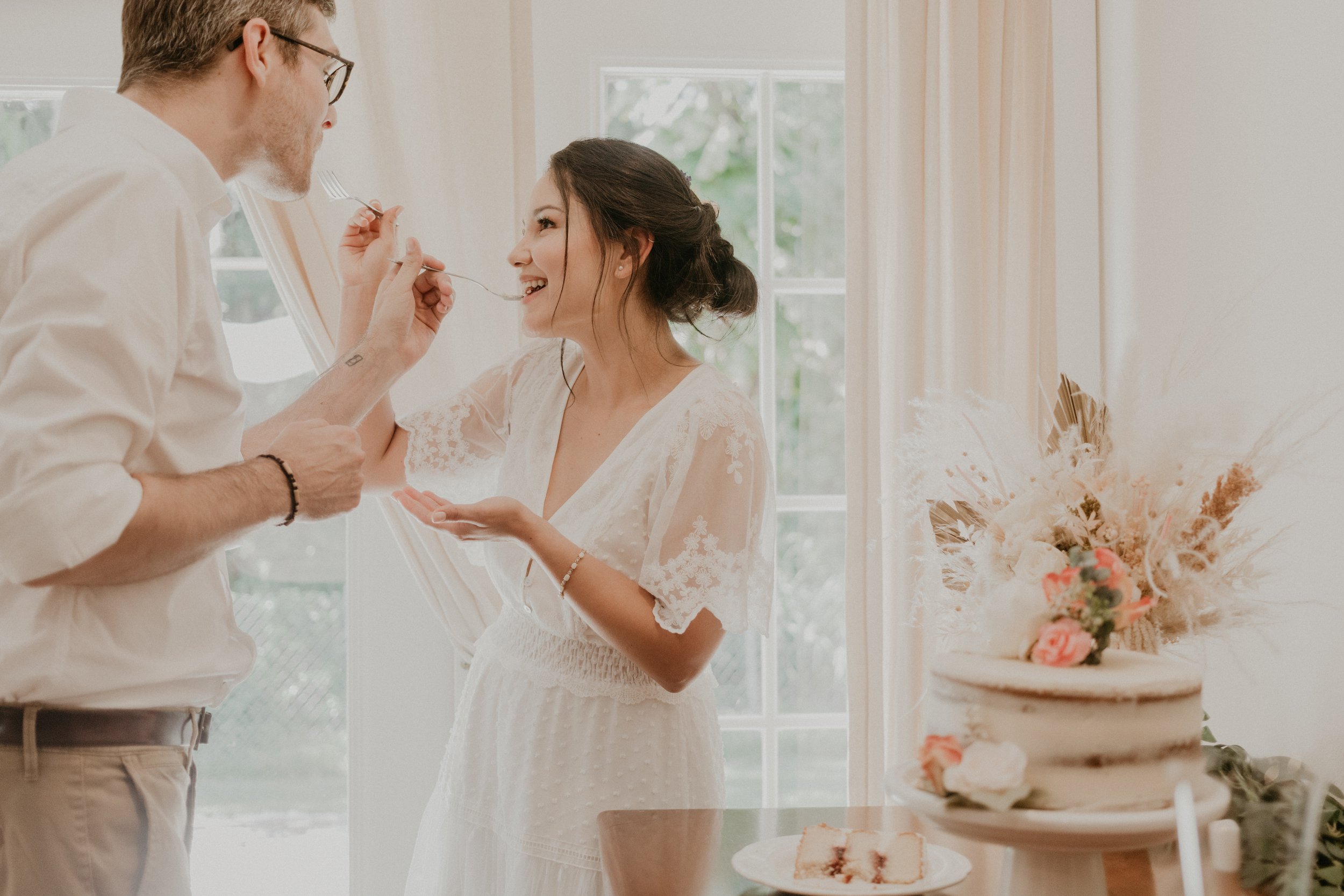 A couple sharing a moment during their elopement celebration, with a woman in a white wedding dress smiling as a man feeds her cake, in a bright room with floral decor and a wedding cake.