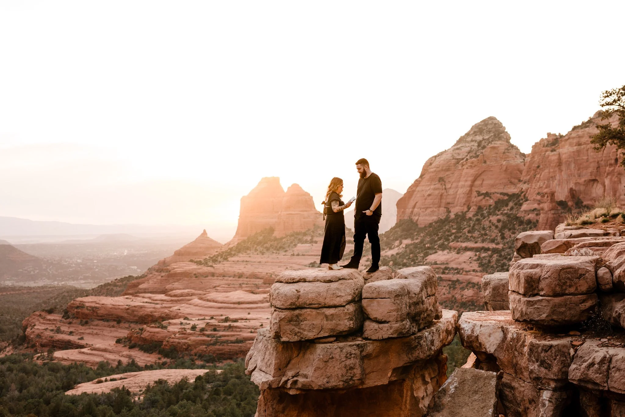 An elopement couple standing on a rock formation overlooking a desert landscape in Sedona, with red rock cliffs and mountains during sunset.