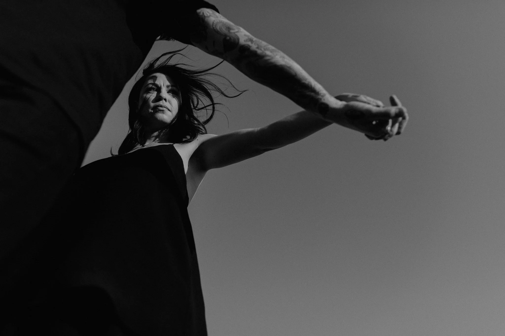 A woman with long dark hair and tattoos on her arm reaches out with her arms extended in a black and white photo taken from a low angle.