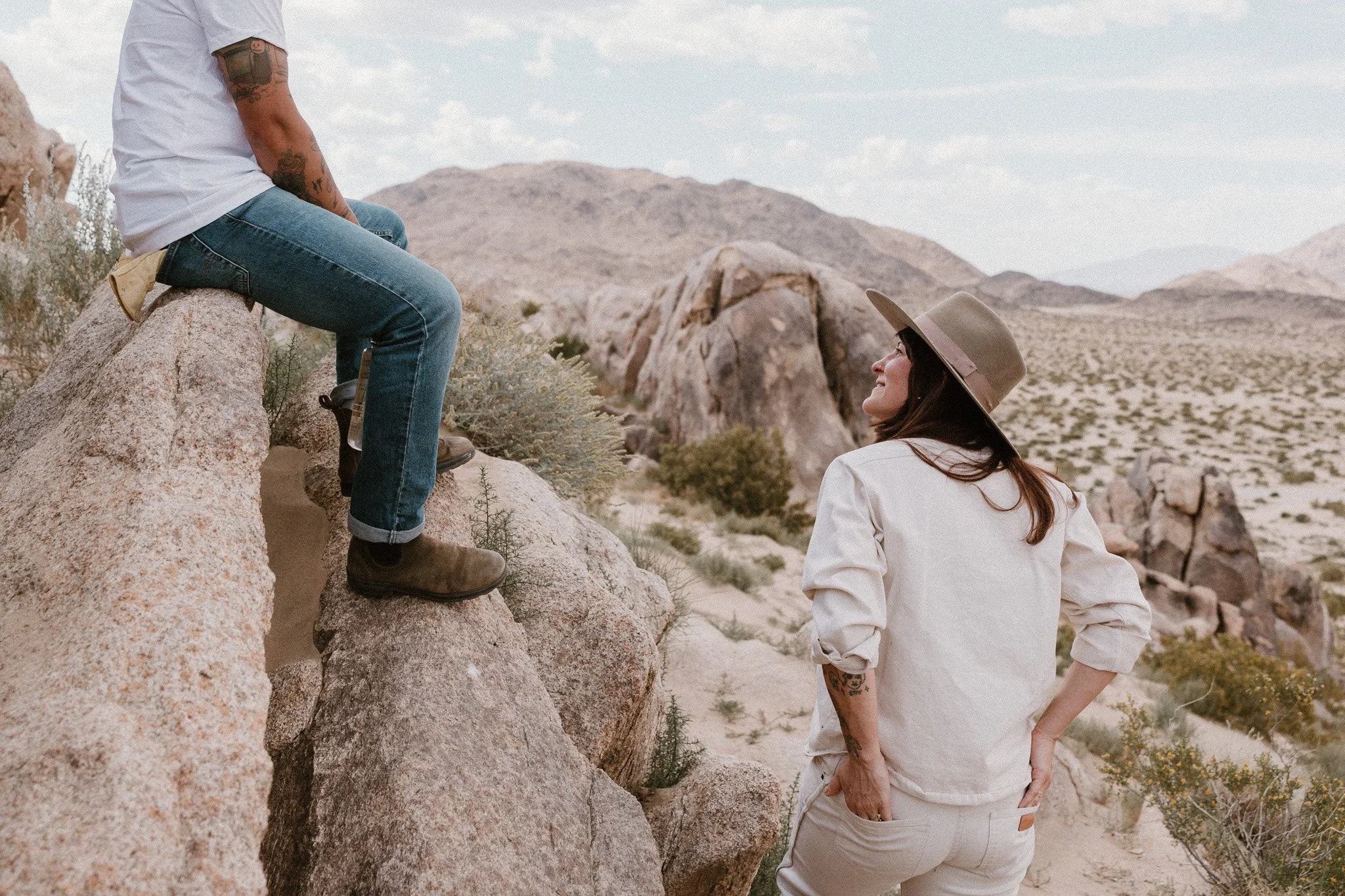 A woman with a hat smiling at a man sitting on a large rock in a desert landscape in Joshua Tree with mountains in the background.