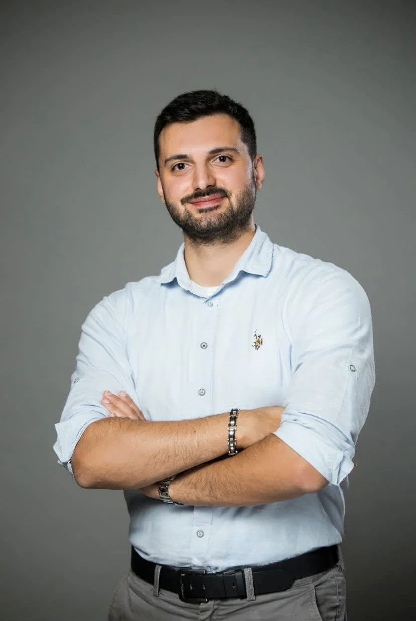 Portret van een man met een korte baard, gekleed in een lichtblauwe overhemd, met de armen over elkaar, poseert voor een grijze achtergrond.