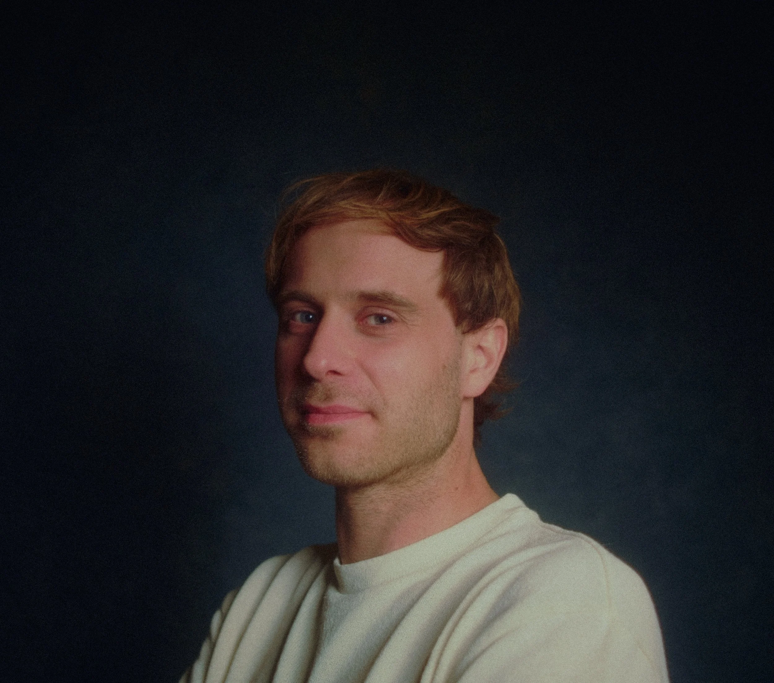Porträt eines Mannes mit braunen, leicht gewellten Haaren, hellblauen Augen und einem leichten Lächeln, trägt ein cremefarbenes Sweatshirt vor dunklem Hintergrund.