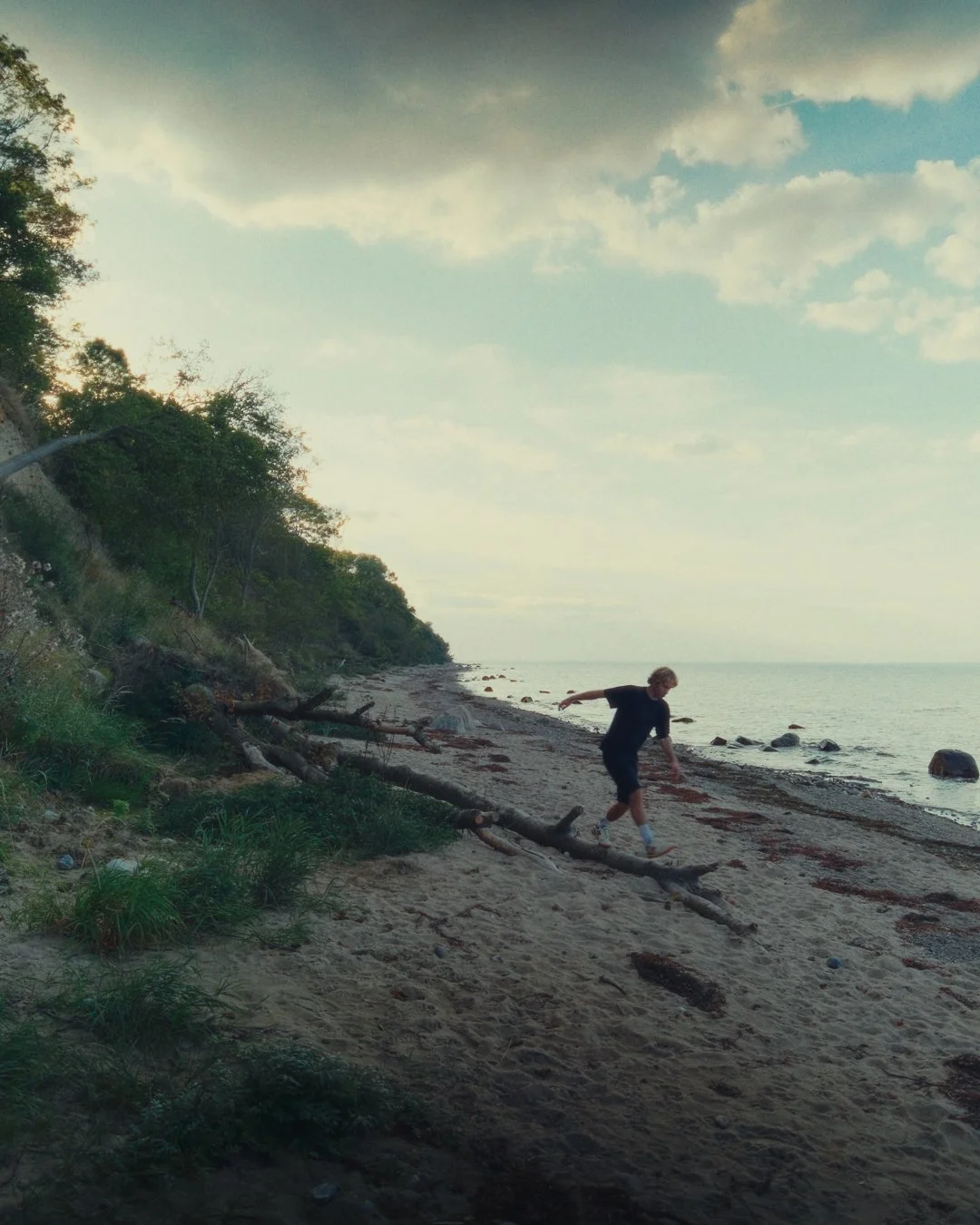 Ein Junge läuft am Strand entlang, mit Wasser und Steinen im Meer im Hintergrund, bei bewölktem Himmel.