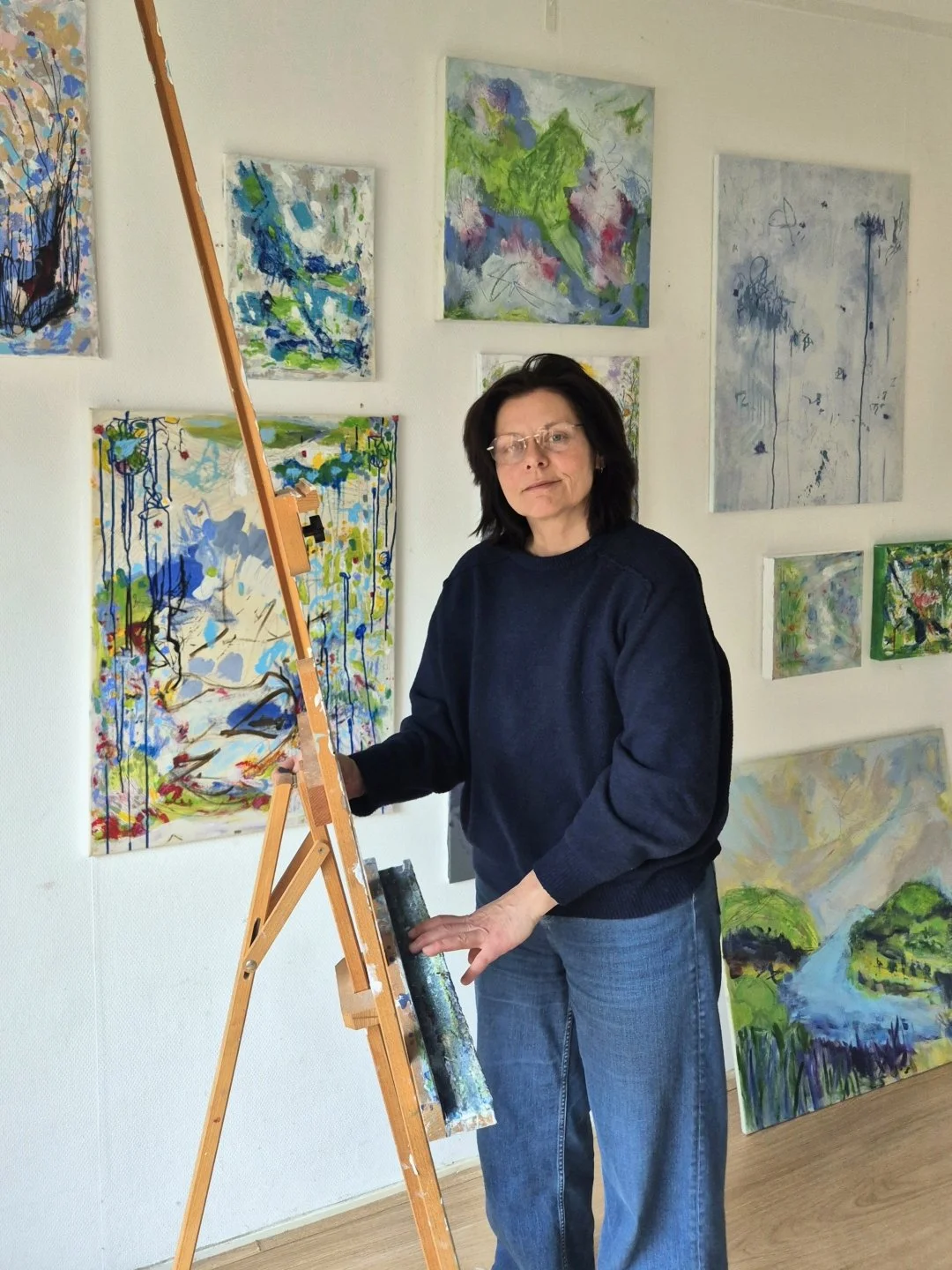 Contemporary abstract artist Daniela Knobel standing in her studio beside an easel, surrounded by layered abstract paintings in blue, green, and soft neutral tones.