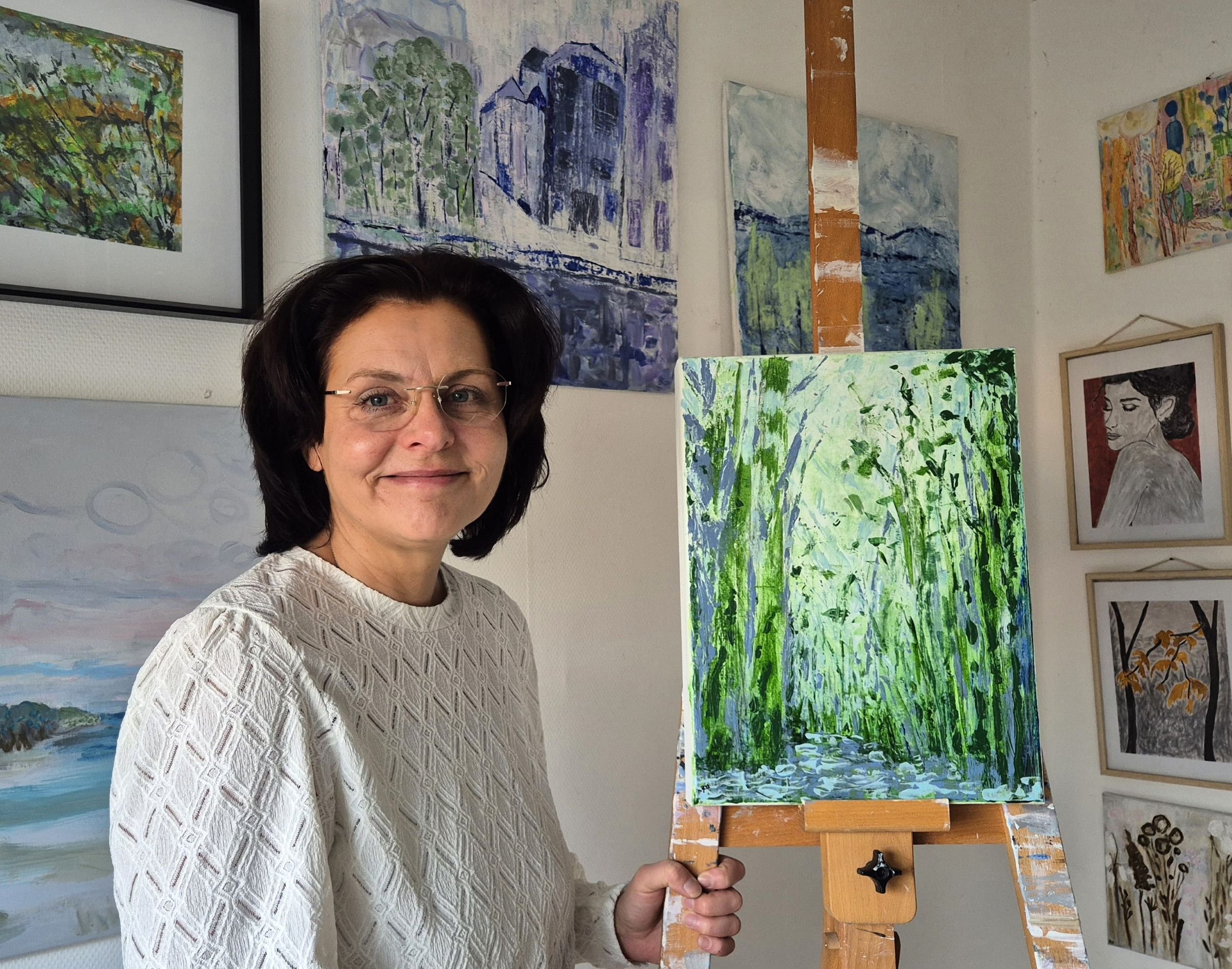 Studio portrait of contemporary abstract artist Daniela Knobel holding a green-toned abstract landscape painting on an easel, in her studio surrounded by paintings.