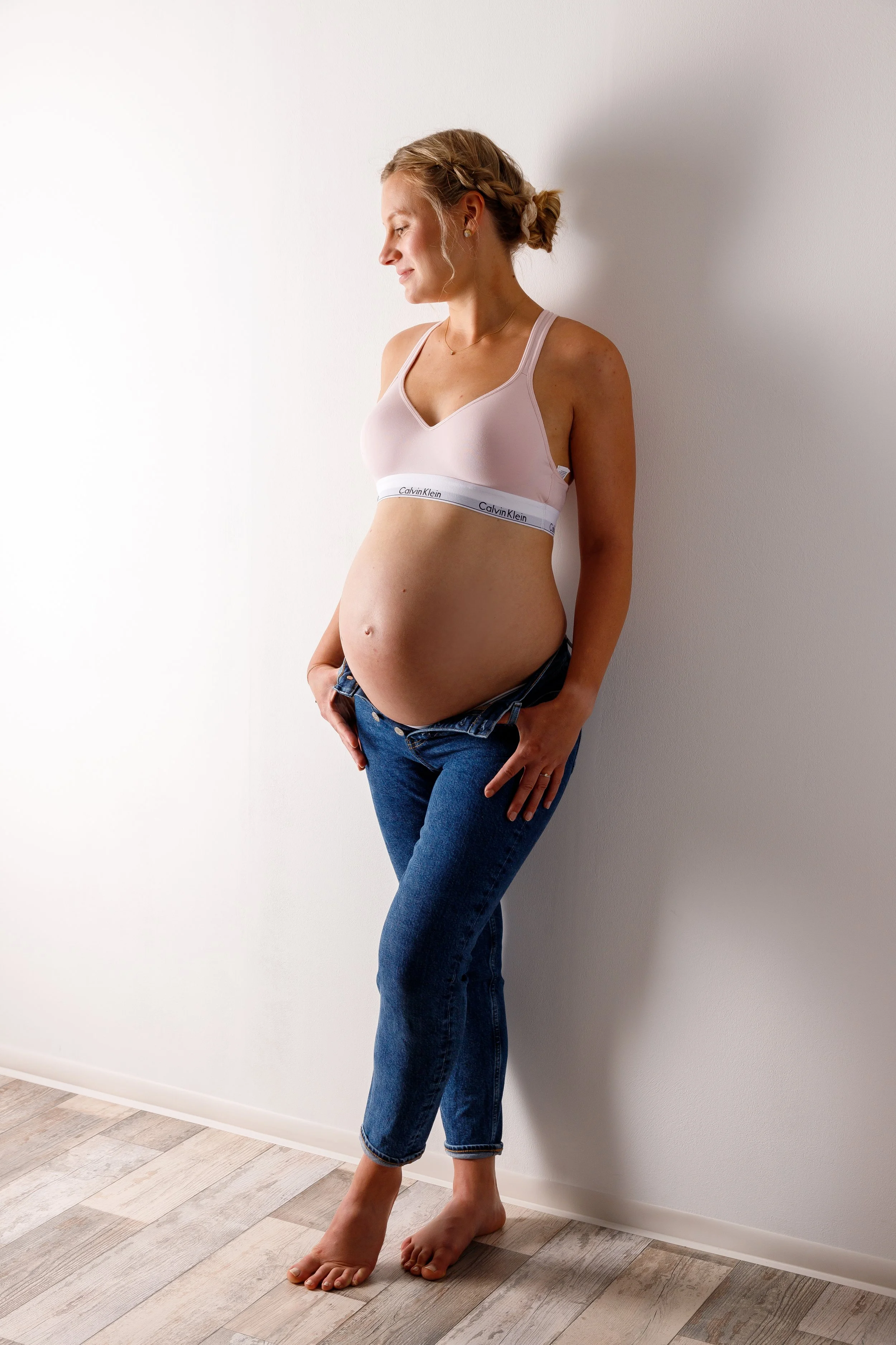 Schwangere Frau in Jeans und rosa BH lehnt an weißer Wand.
