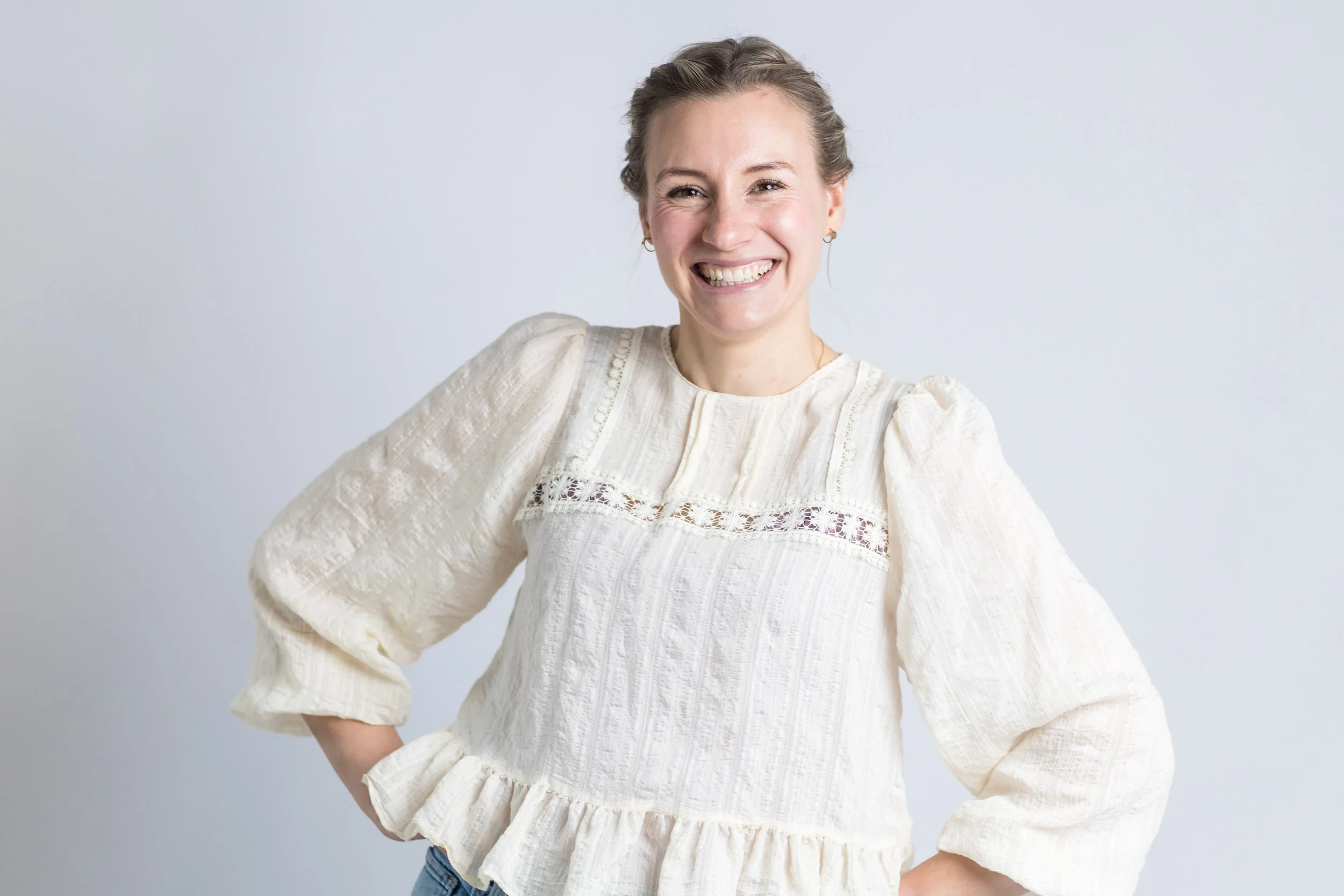 Lächelnde Mama mit lockigem Haar in einem cremefarbenen, dekorativen Oberteil vor hellem Hintergrund.