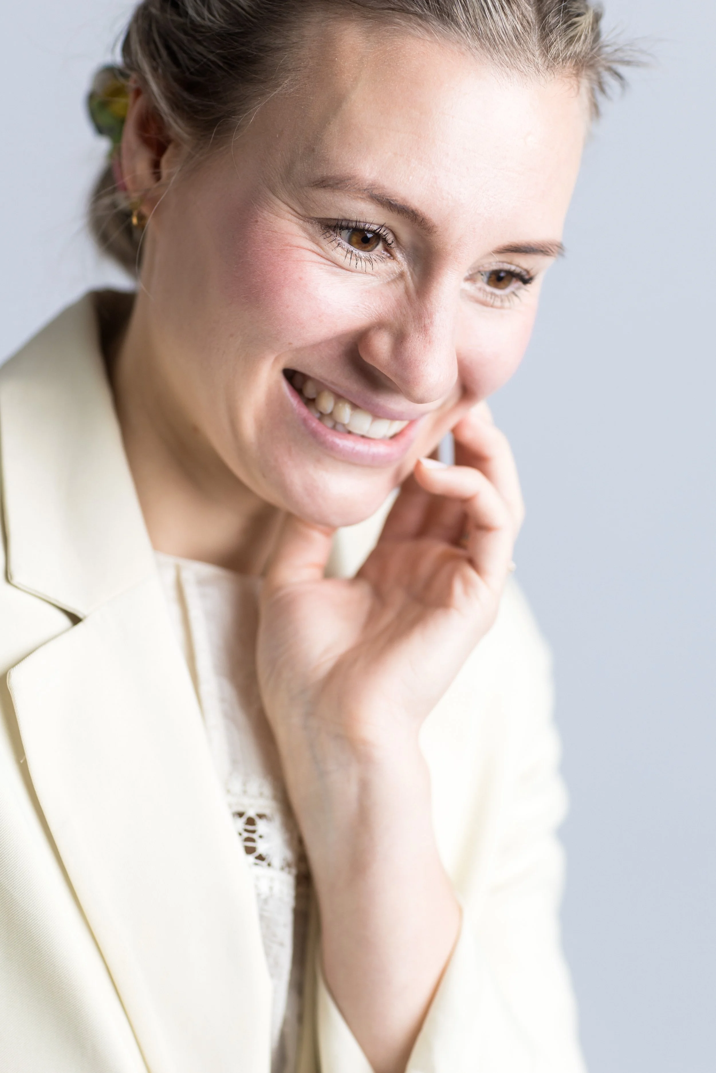 Nahaufnahme eines lachenden, jungen Frau mit braunen Haaren, die ihre Hand an ihr Gesicht legt, gegen einen hellen Hintergrund.