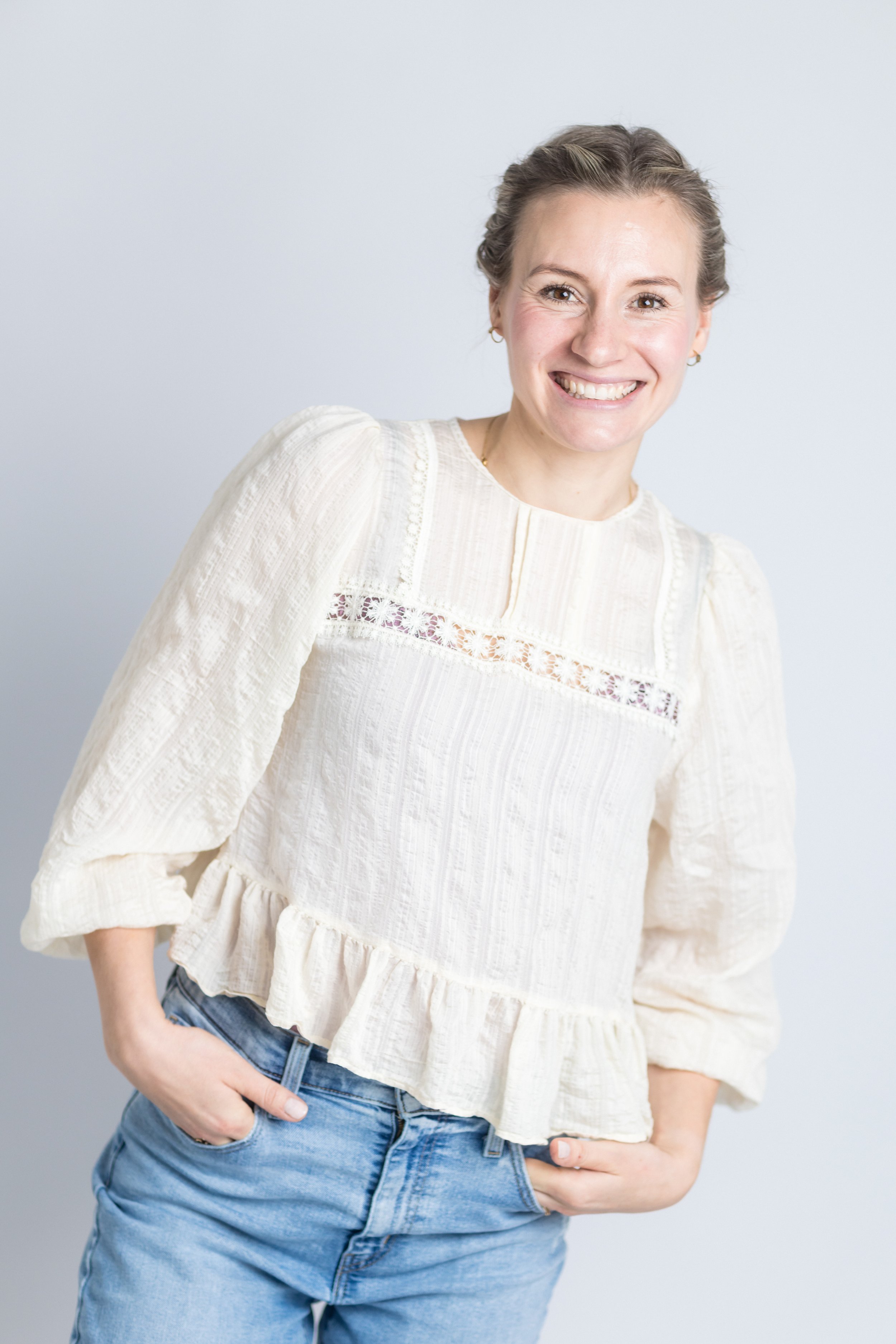 Junge Frau mit braunen Haaren und weißem Spitzenbluse lächelnd vor hellem Hintergrund.