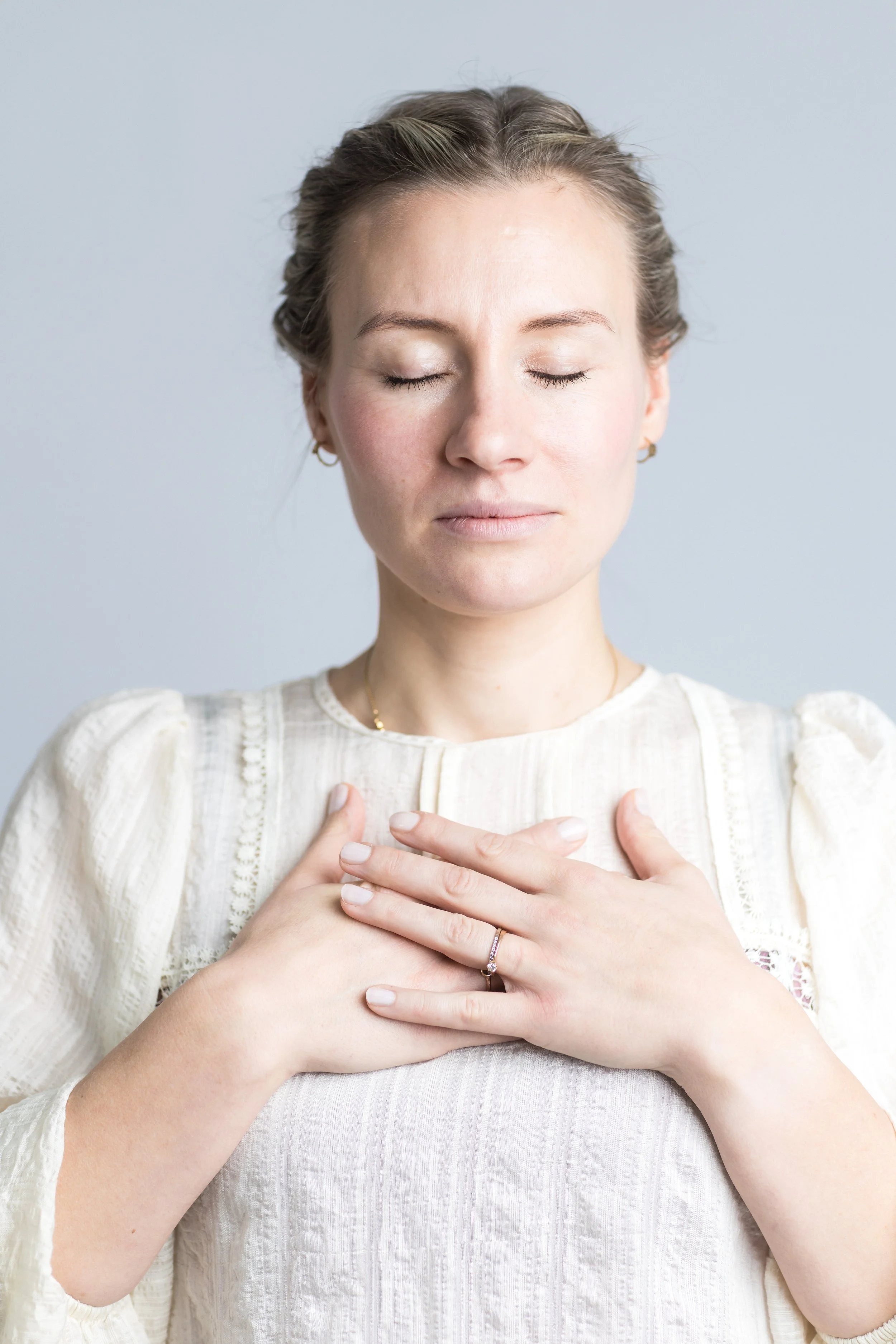 Eine Mama mit geschlossenen Augen legt eine Hand auf die Brust, trägt einen Ring und eine goldene Halskette, vor einem hellblauen Hintergrund.