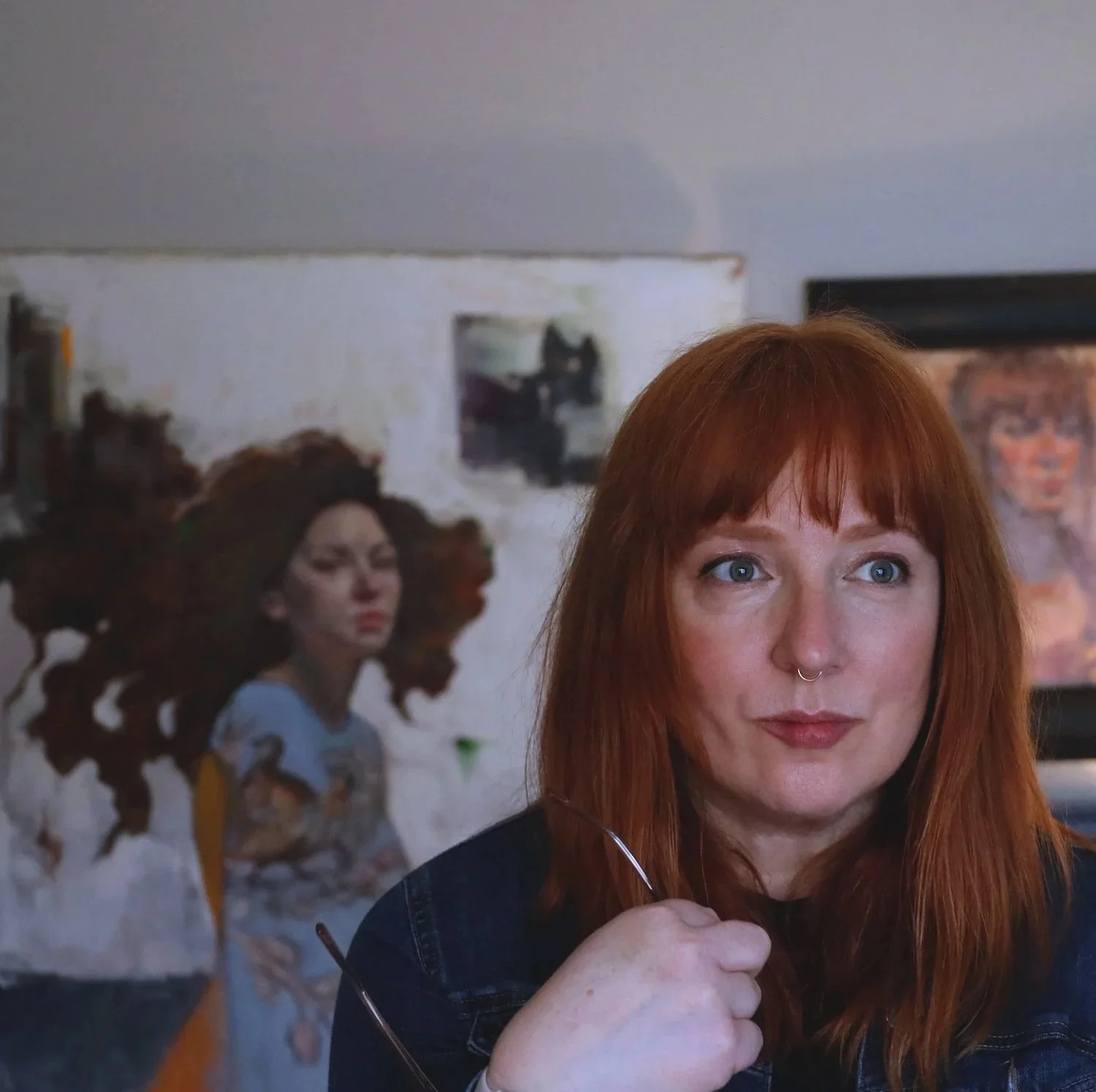 A woman with red hair and a septum piercing is sitting in front of a wall with artwork, holding glasses in her hand. Behind her, there is a painting of a woman with curly hair and a portrait of a woman on a screen.