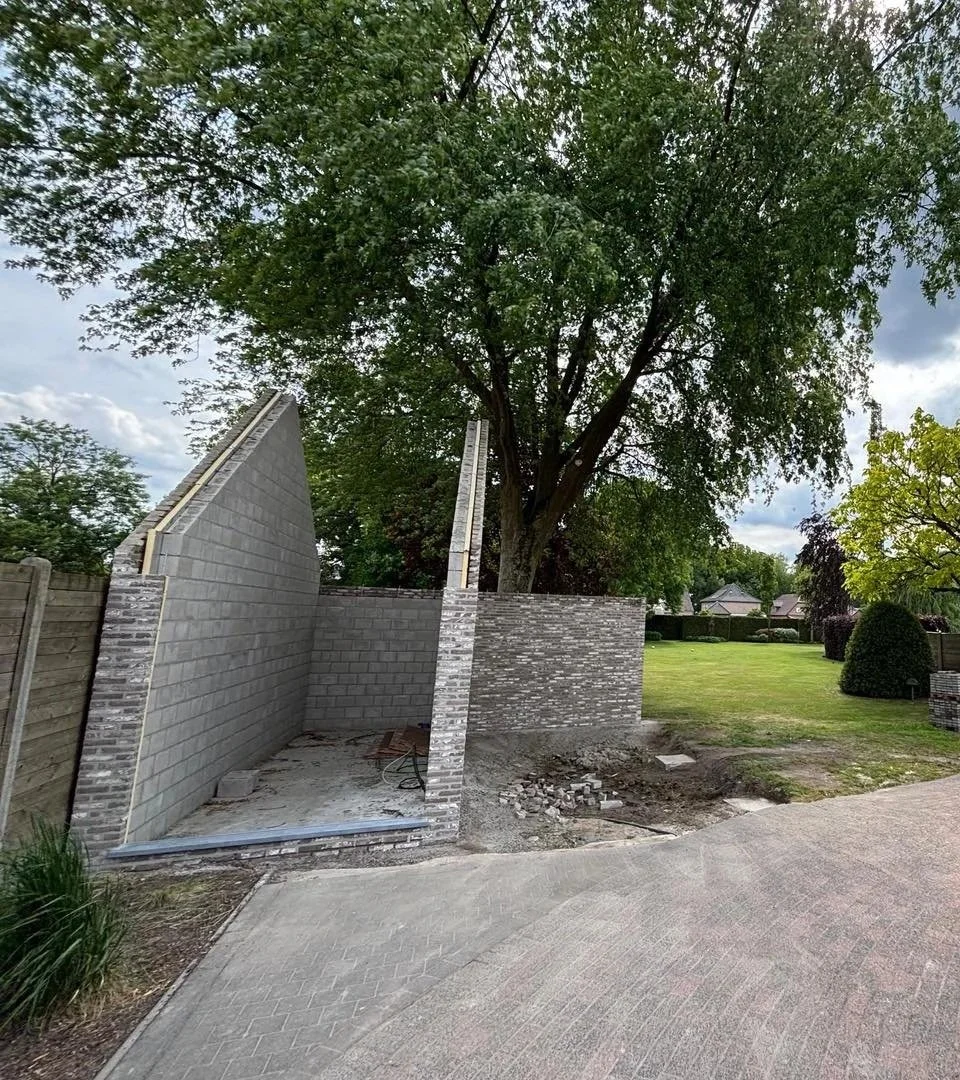 Een tuin met een bestaande boom, een grassveld en een oprit, met een bouwproject voor een nieuwe muur of kamer onder constructie, waarbij bakstenen en cement worden gebruikt voor een carport.